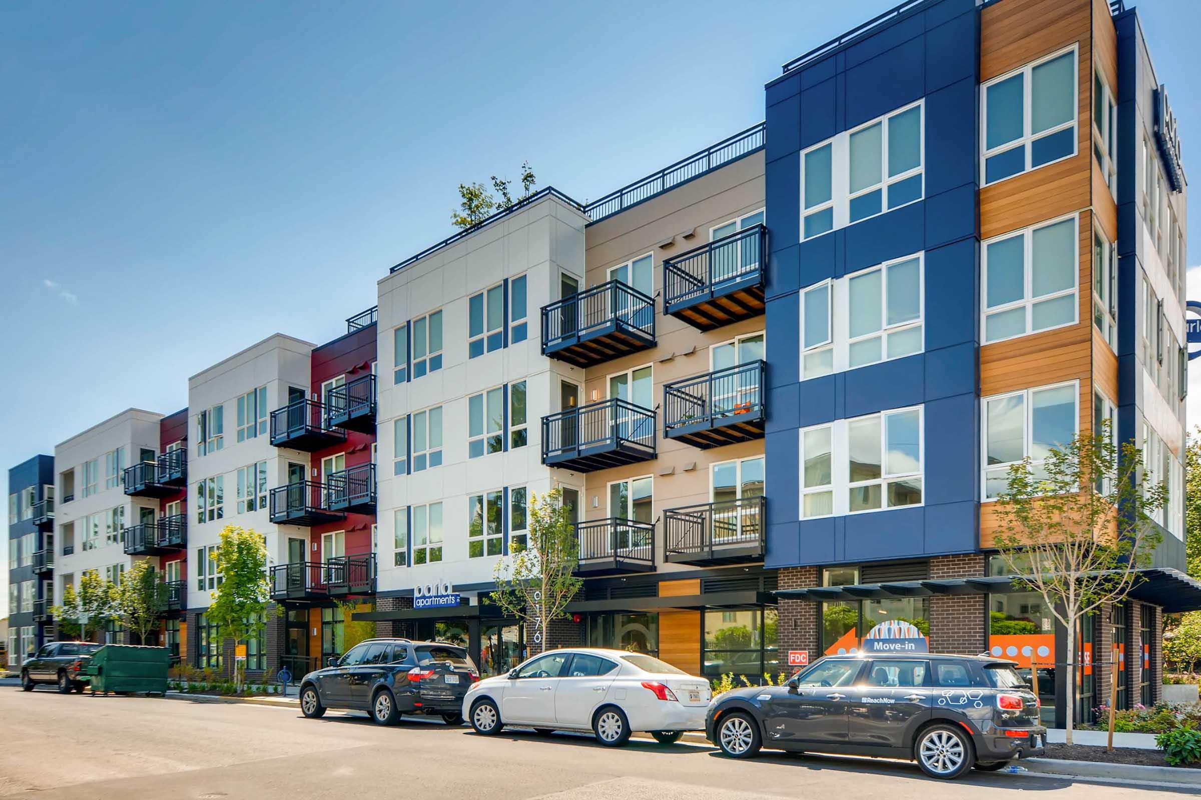 Modern apartment buildings with a mix of white, red, and blue facades, featuring balconies and large windows. The street is lined with cars and small trees, with a storefront visible on the ground level. Clear blue sky in the background.