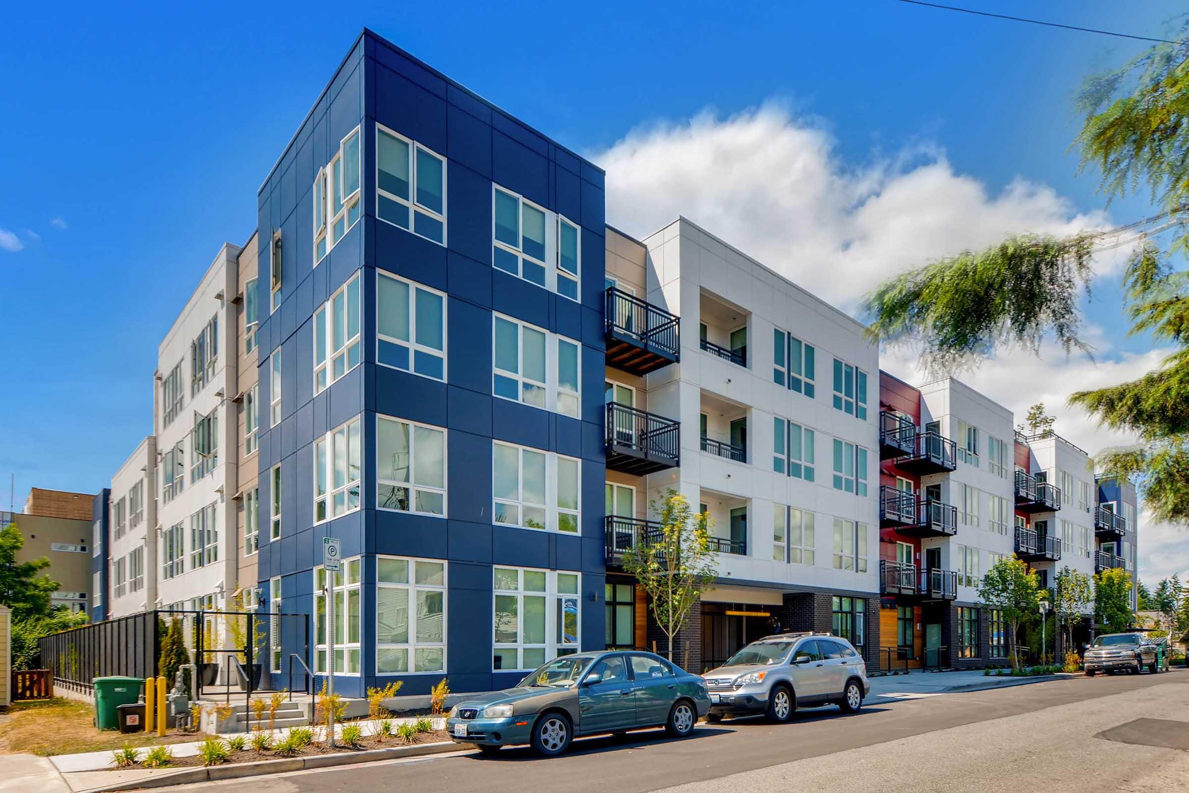 Modern apartment building featuring a mix of blue and white exteriors with large windows. Balconies are visible on several floors, and cars are parked along the street in front. Trees and landscaping enhance the urban setting under a partly cloudy sky.
