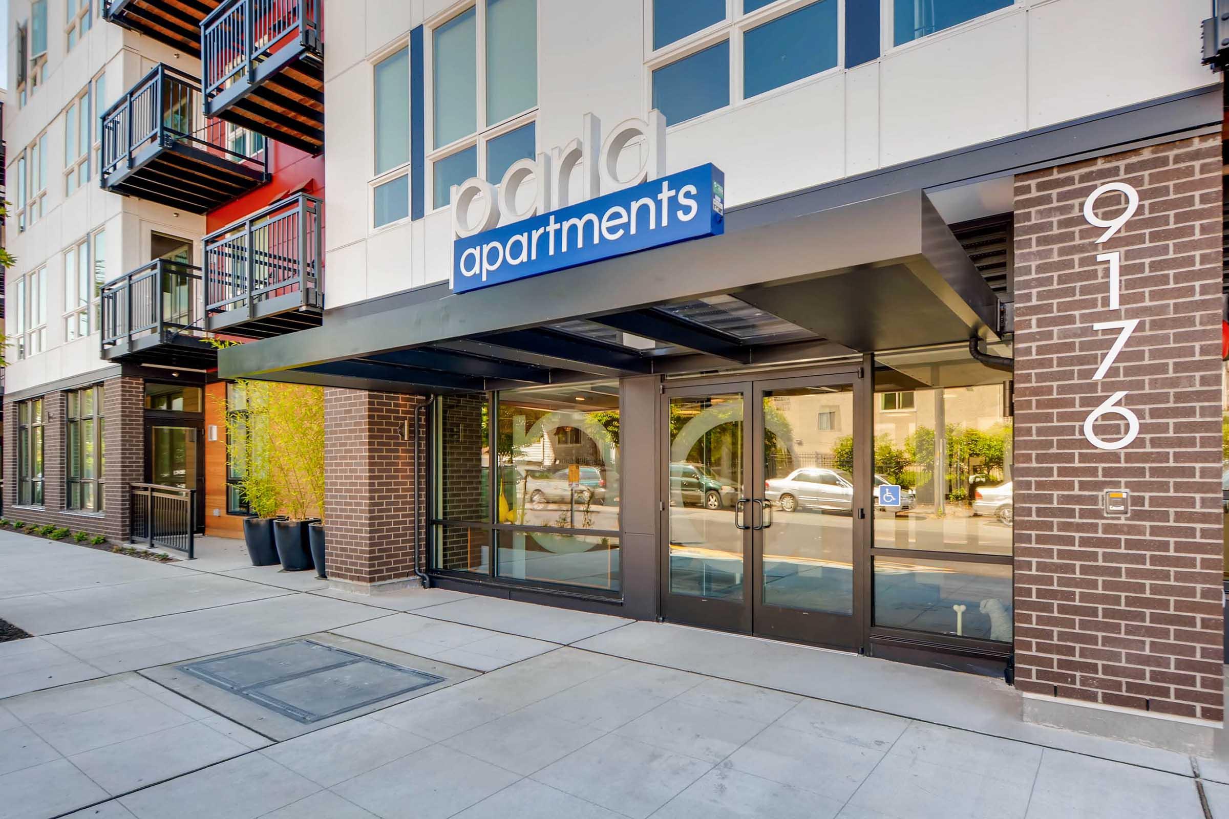 Entrance of a modern apartment building featuring the sign "parla apartments." The building has a brick facade, large glass doors, and balconies. The address number "9176" is displayed on the right side. Potted plants are visible near the entrance, enhancing the inviting atmosphere.