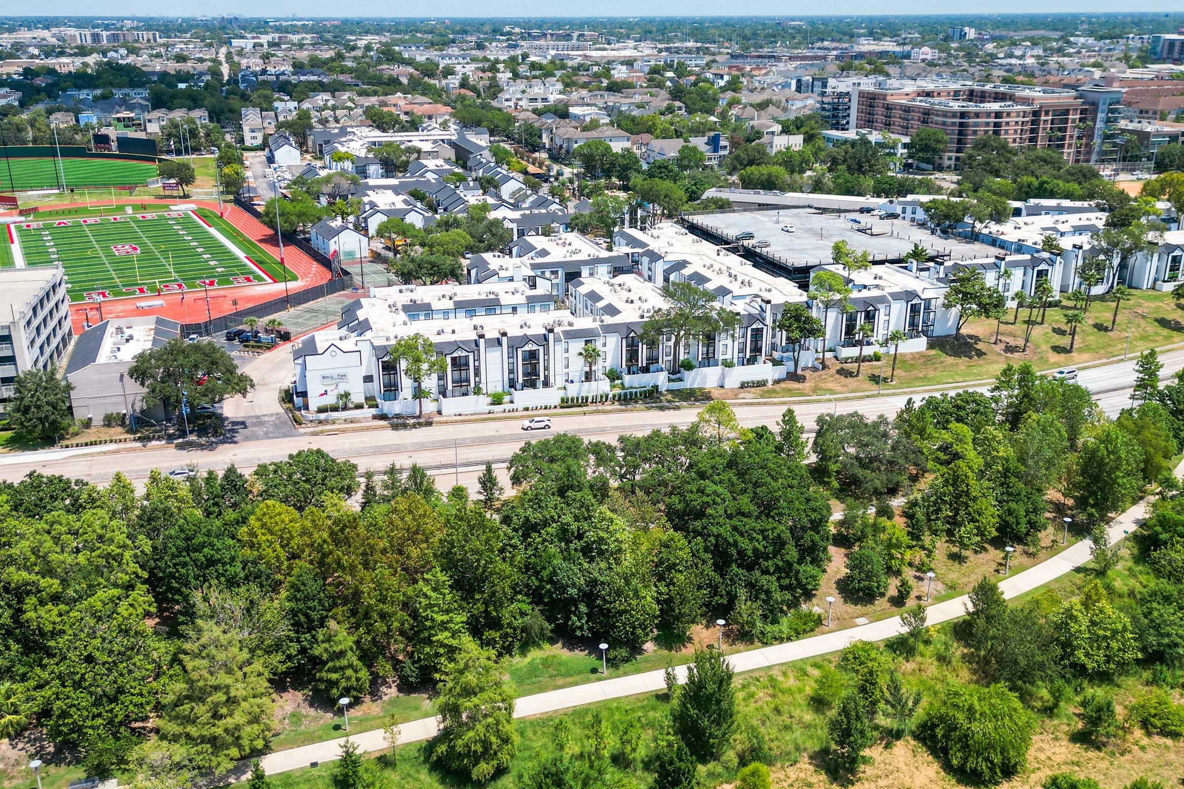 Aerial view of an urban area featuring residential buildings, green spaces, and a sports field. The sports field has a red track and bright green turf. Surrounding the area are trees and pathways, with a mix of residential and commercial structures visible in the background.