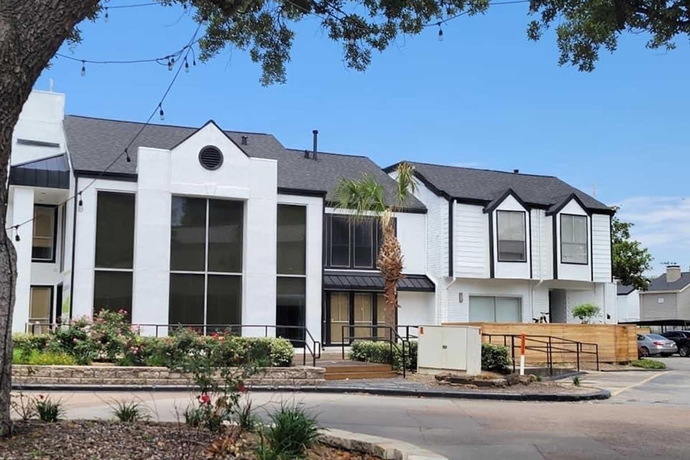 Modern multi-story residential building featuring a mix of white stucco and dark accents. Large windows on the ground floor allow natural light, with palm trees and landscaping in the foreground. A wooden fence and a pathway are visible, complementing the contemporary architecture against a clear blue sky.