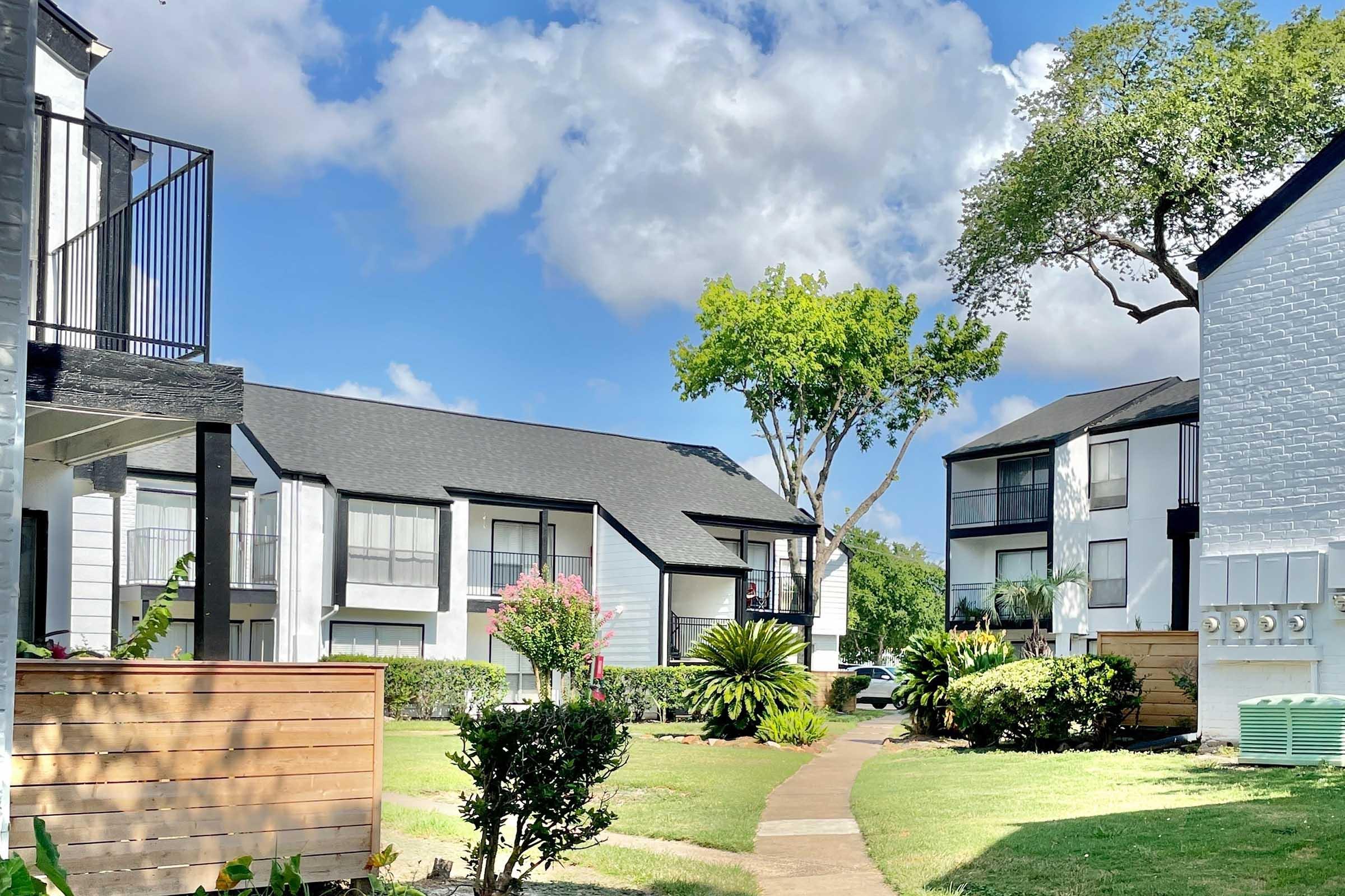 A serene residential area featuring two-story apartment buildings with a mix of greenery, including palm trees and flowering plants. A pathway leads through the landscaped yard under a bright blue sky with fluffy clouds, creating a peaceful atmosphere.