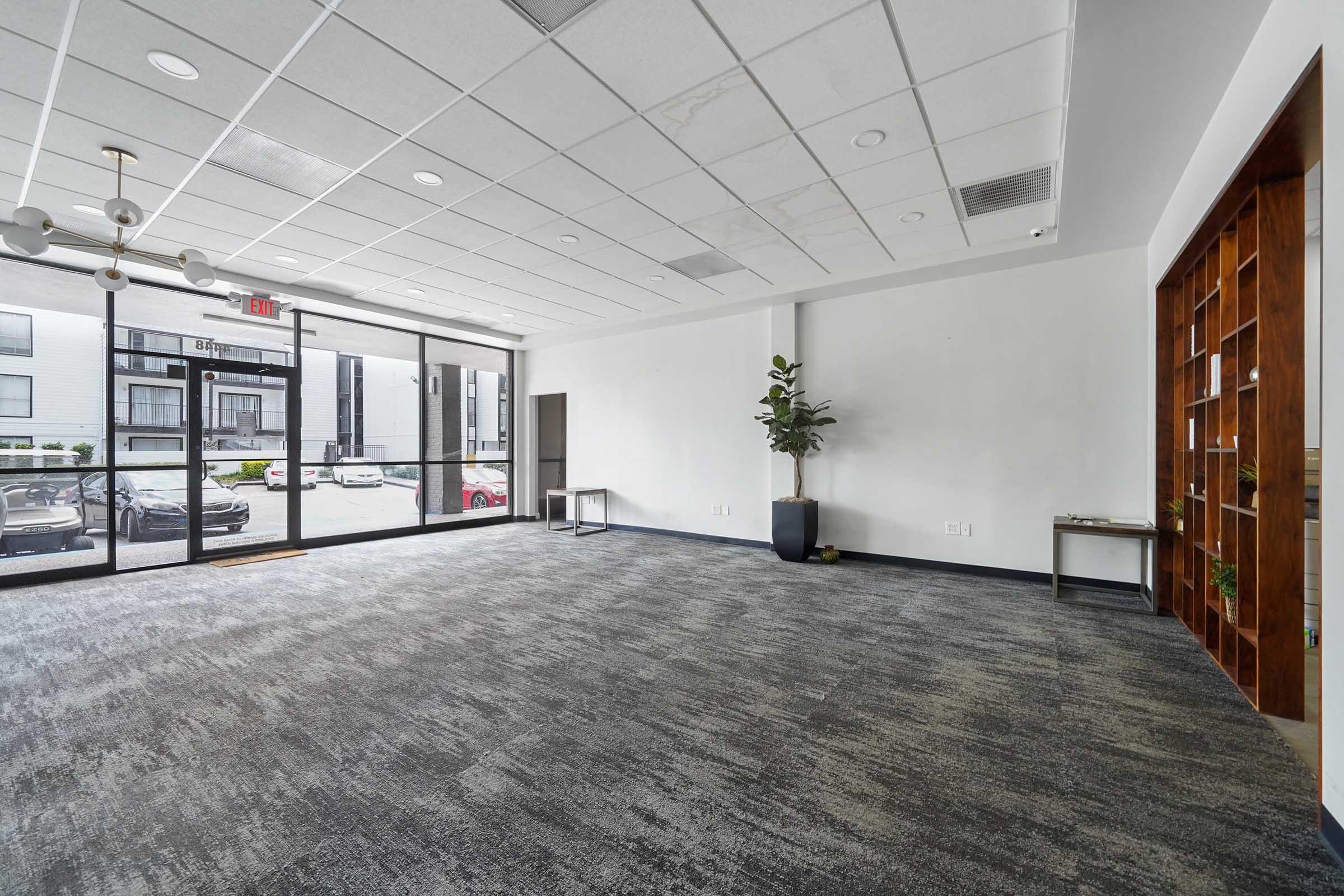 A spacious, modern lobby area with a gray carpet, large glass doors allowing natural light, and minimalistic decor. A green plant is placed in a corner, and there's a simple table against the wall. A wooden bookshelf is visible, showcasing a few decorative items. The overall atmosphere is clean and inviting.