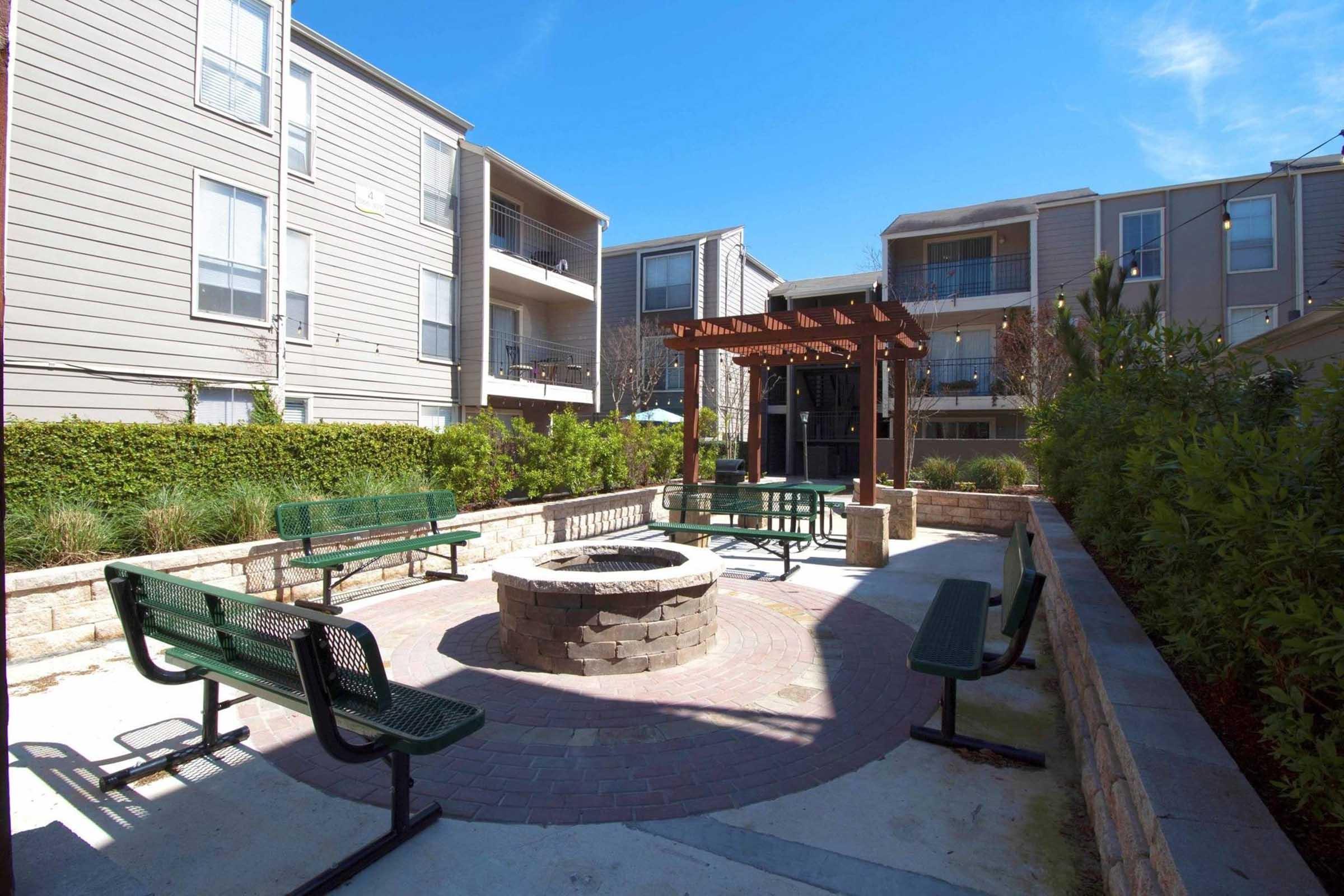 A communal outdoor space featuring a circular stone fire pit surrounded by benches, shaded by a wooden pergola. The area is landscaped with greenery and is flanked by multi-story residential buildings under a clear blue sky.