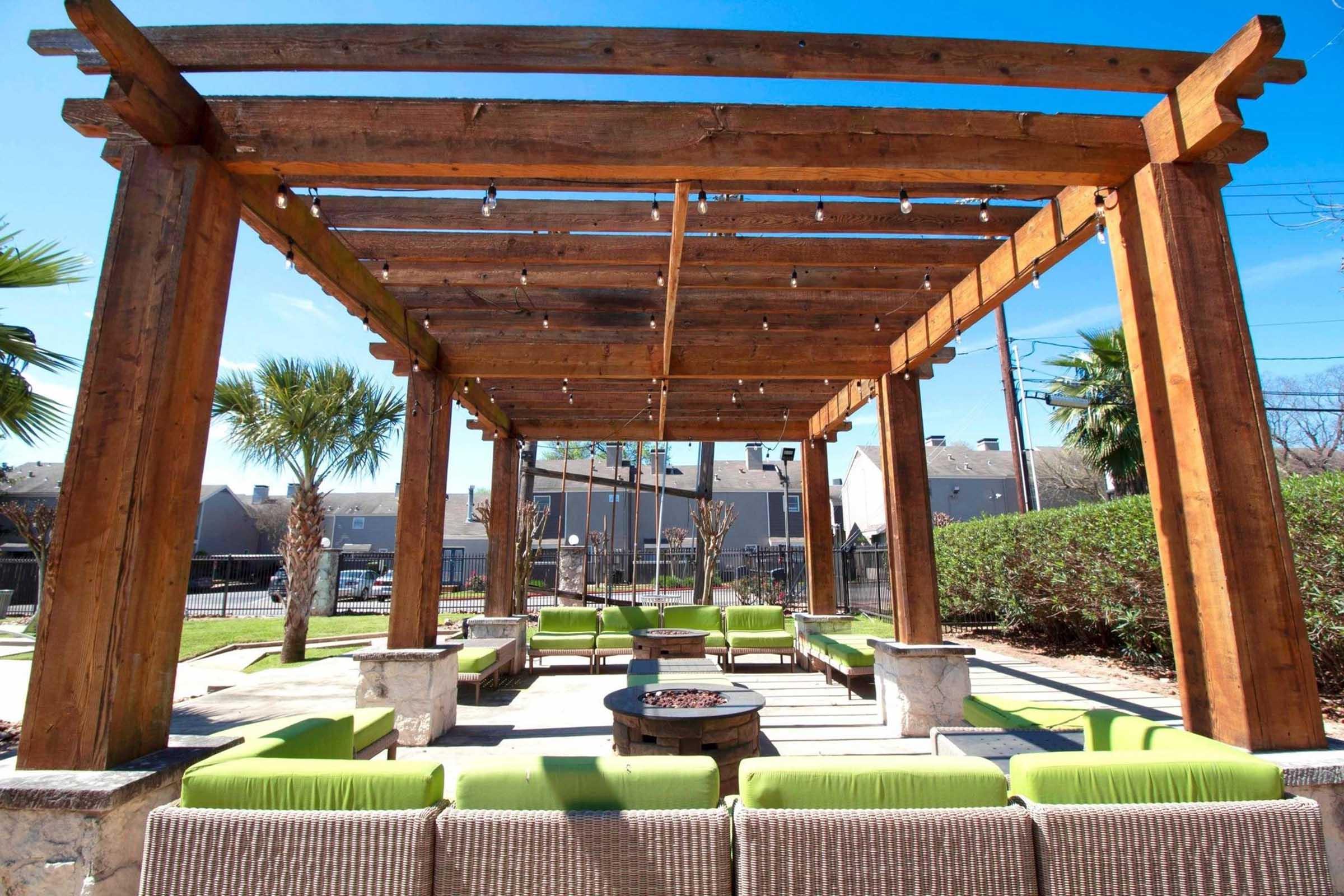 A spacious outdoor seating area under a wooden pergola with string lights, featuring green lounge chairs arranged around a circular fire pit. Lush landscaping and palm trees provide a tropical feel in a sunny environment, creating a relaxing space for gatherings.