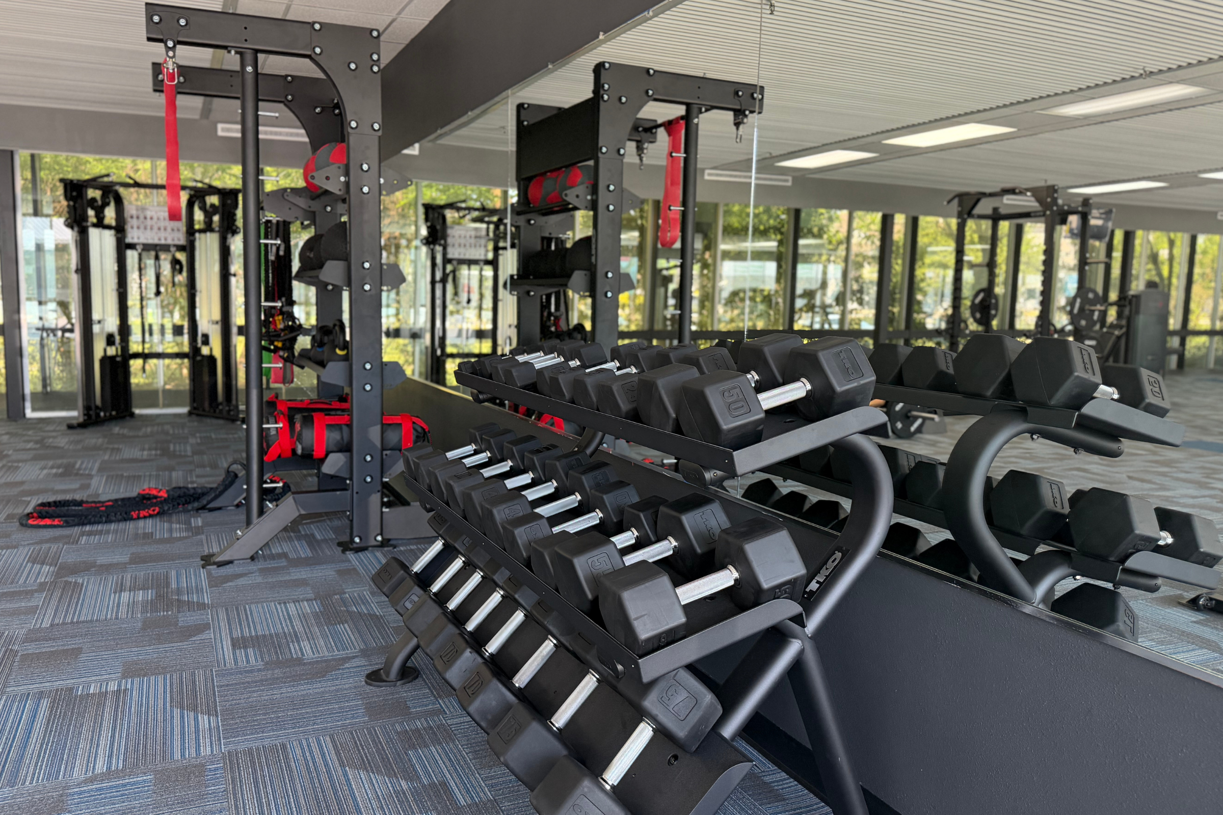 A modern gym interior featuring a rack of dumbbells in the foreground, with weightlifting stations and fitness equipment visible in the background. The space is well-lit with large windows and mirrors, creating an open and inviting workout environment.