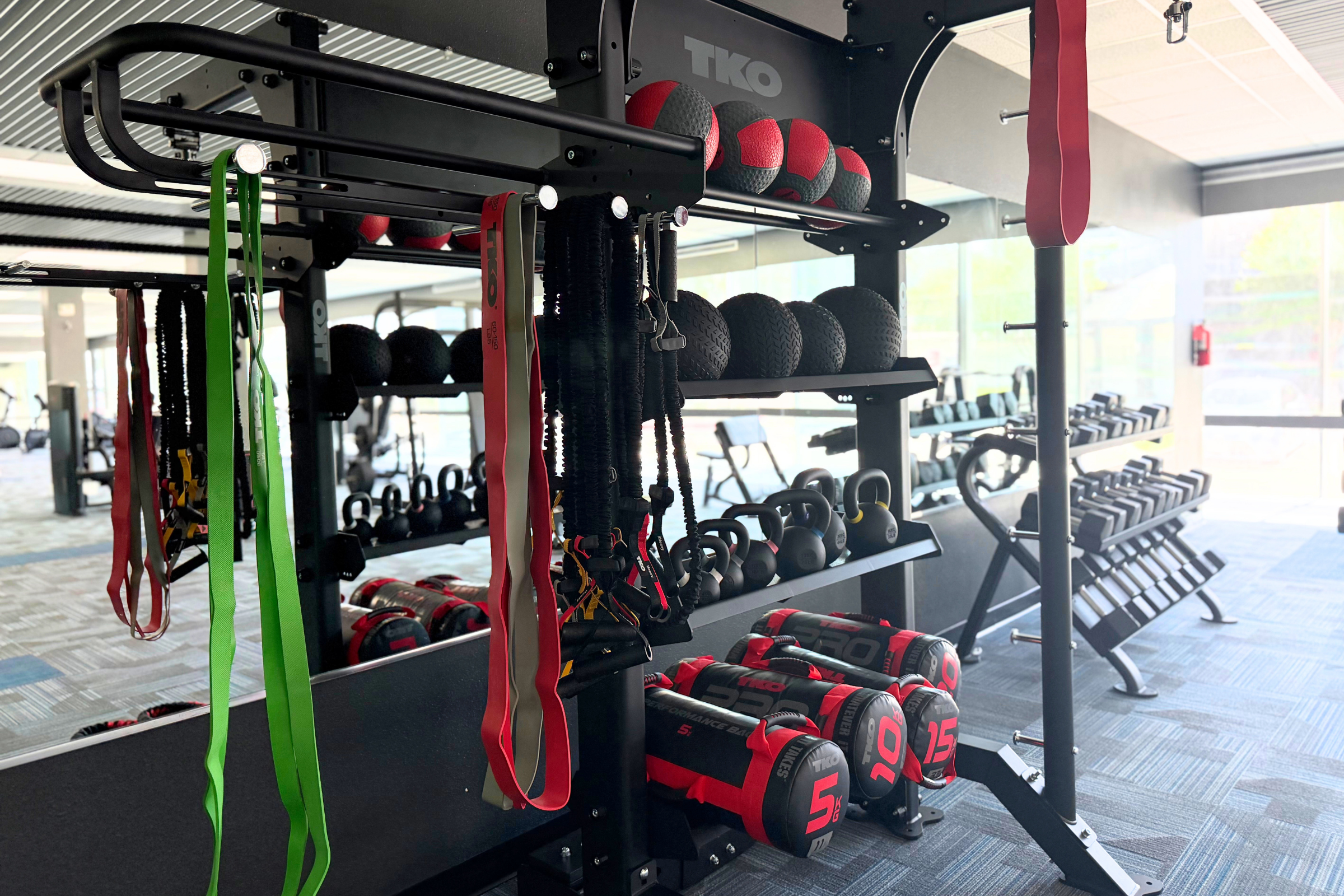 A fitness facility featuring various workout equipment, including kettlebells, resistance bands in green and red, and a rack of weighted balls. The area has a mirrored wall, and the ambiance is bright with natural light.
