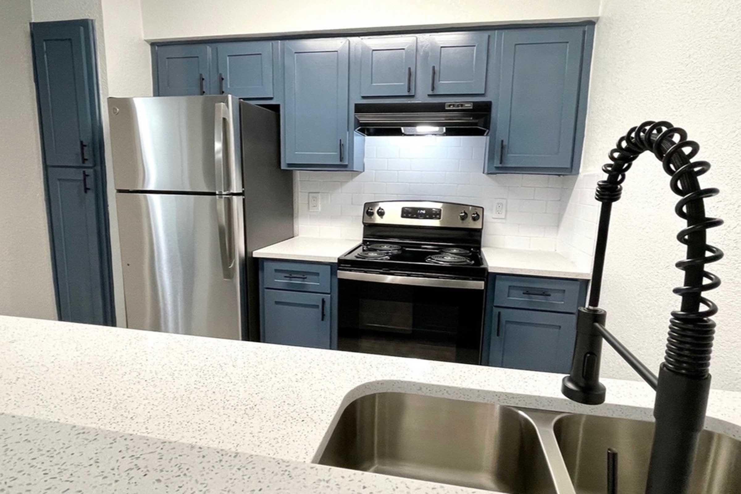 A modern kitchen featuring blue cabinetry, a stainless steel refrigerator, an oven with a hood, and a black stovetop. There is a white countertop with a sink and a stylish faucet, all set in a well-lit, contemporary space. The overall design is clean and functional.