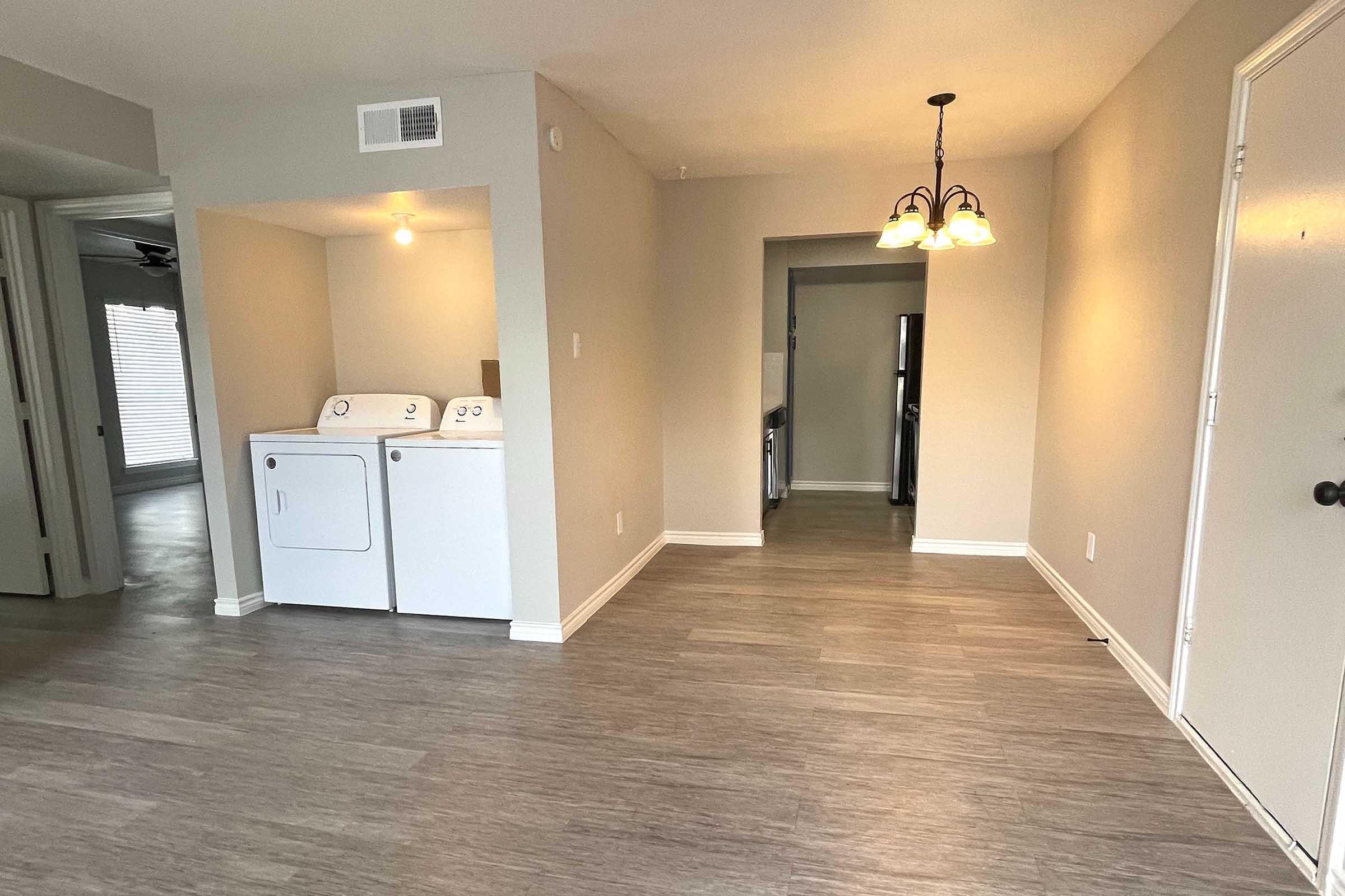 A view of a light-filled apartment interior featuring a laundry area with stacked washer and dryer on the left. The flooring is a modern laminate in neutral tones, and there’s a doorway leading to another room in the background. A small chandelier adds a cozy touch to the space.