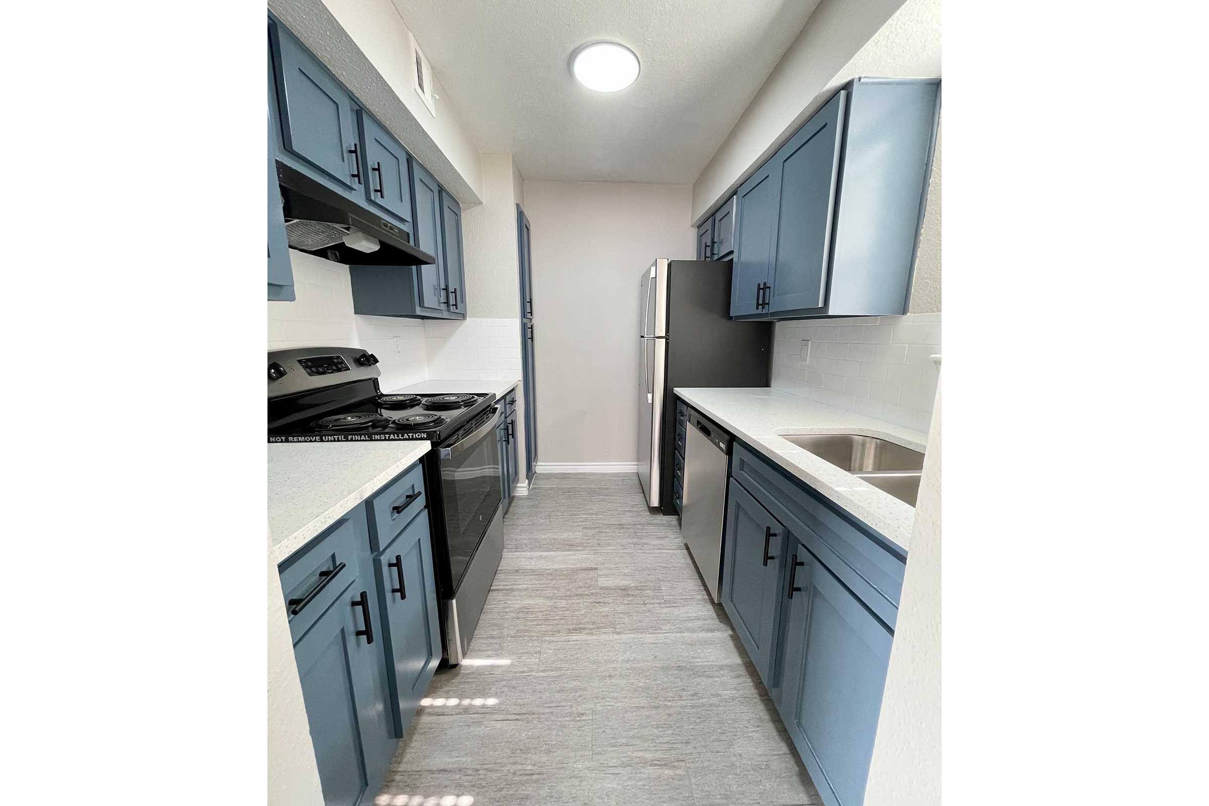 A modern kitchen featuring blue cabinets, a stainless steel refrigerator, and a gas stove. The space has light-colored countertops and a double sink, with natural light coming from a circular ceiling fixture. The walls are painted a soft light color, enhancing the airy feel of the room.