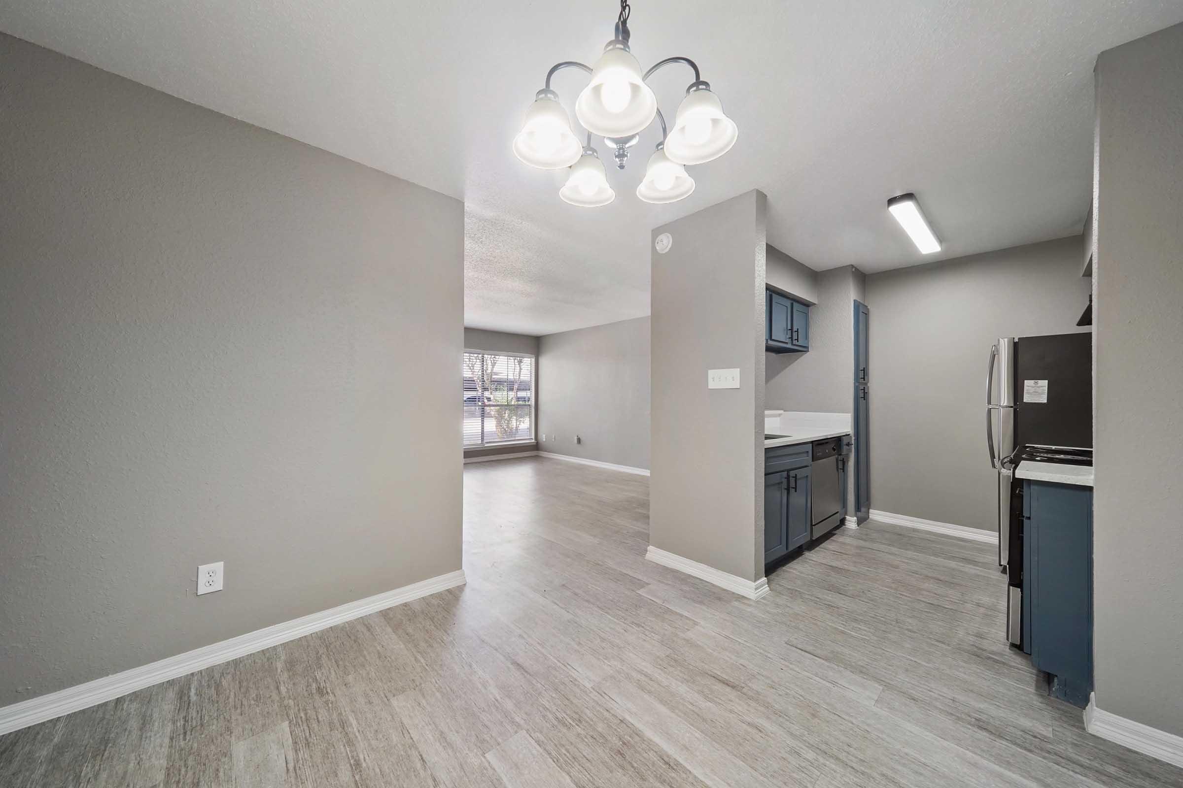 A spacious and modern kitchen area features light gray walls, a stylish chandelier with five bulbs, and blue cabinetry. The living area beyond has large windows providing natural light, and the flooring is a light wood-like laminate, creating a warm and inviting atmosphere.