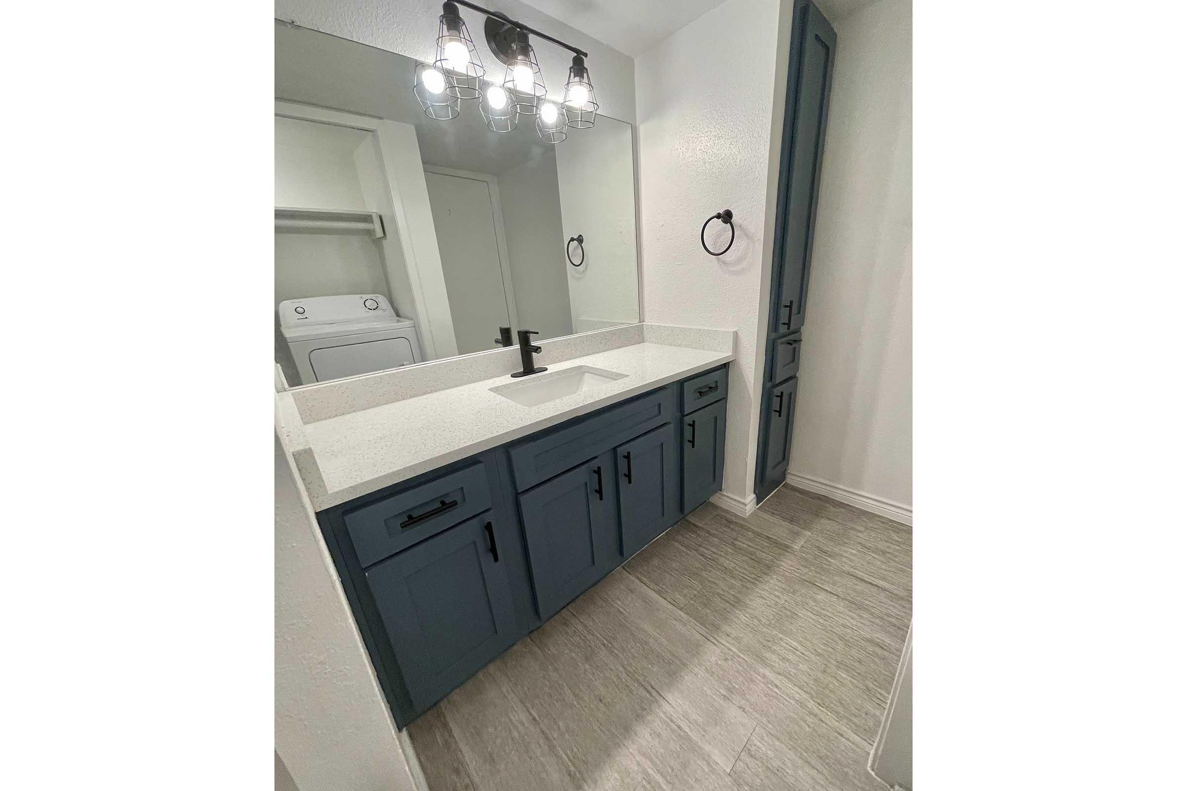 A modern bathroom featuring a navy blue vanity with multiple drawers, a white countertop, and a sleek black faucet. Illuminated by stylish pendant lights overhead, the space includes a large mirror and a wall-mounted towel ring. A laundry area is visible in the background, enhancing the room's functionality.