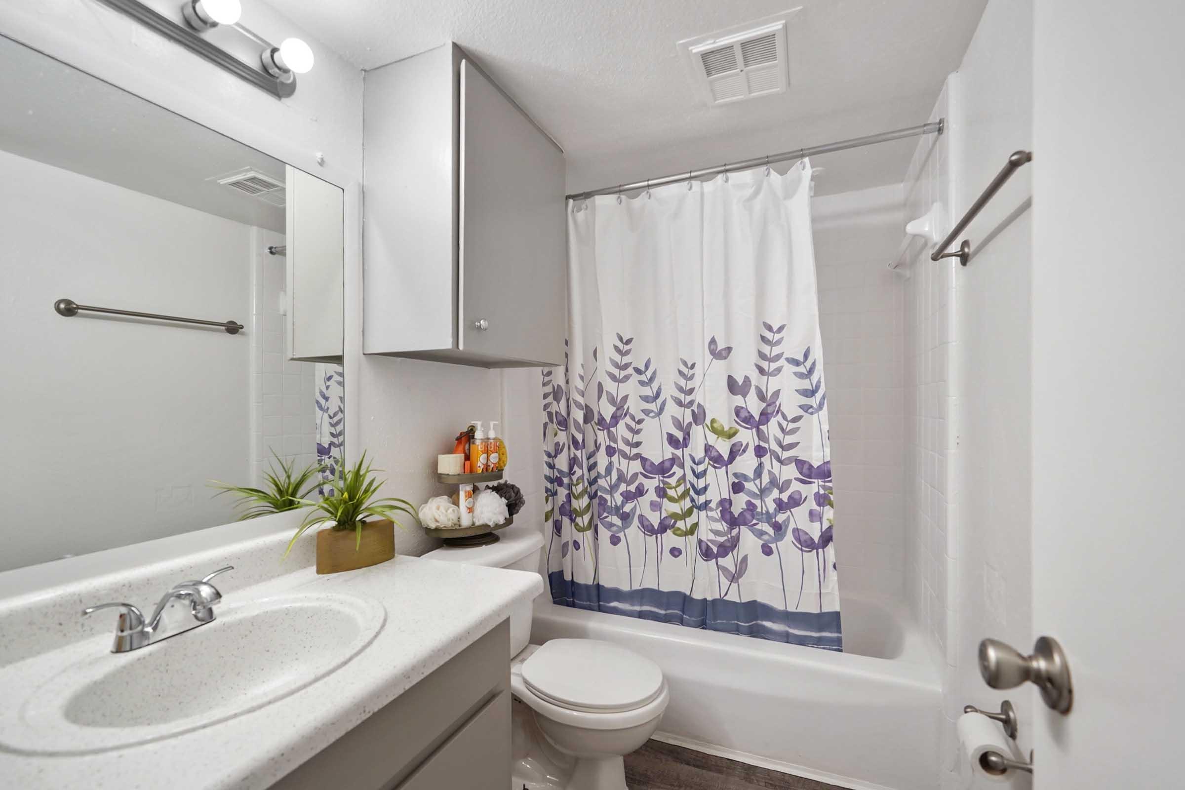 A modern bathroom featuring a white bathtub with a floral shower curtain, a white countertop with a sink, and a mirror above it. The space includes a towel rack, cabinetry, and decorative items like a small plant and toiletries on display. Light fixtures are mounted above the mirror.