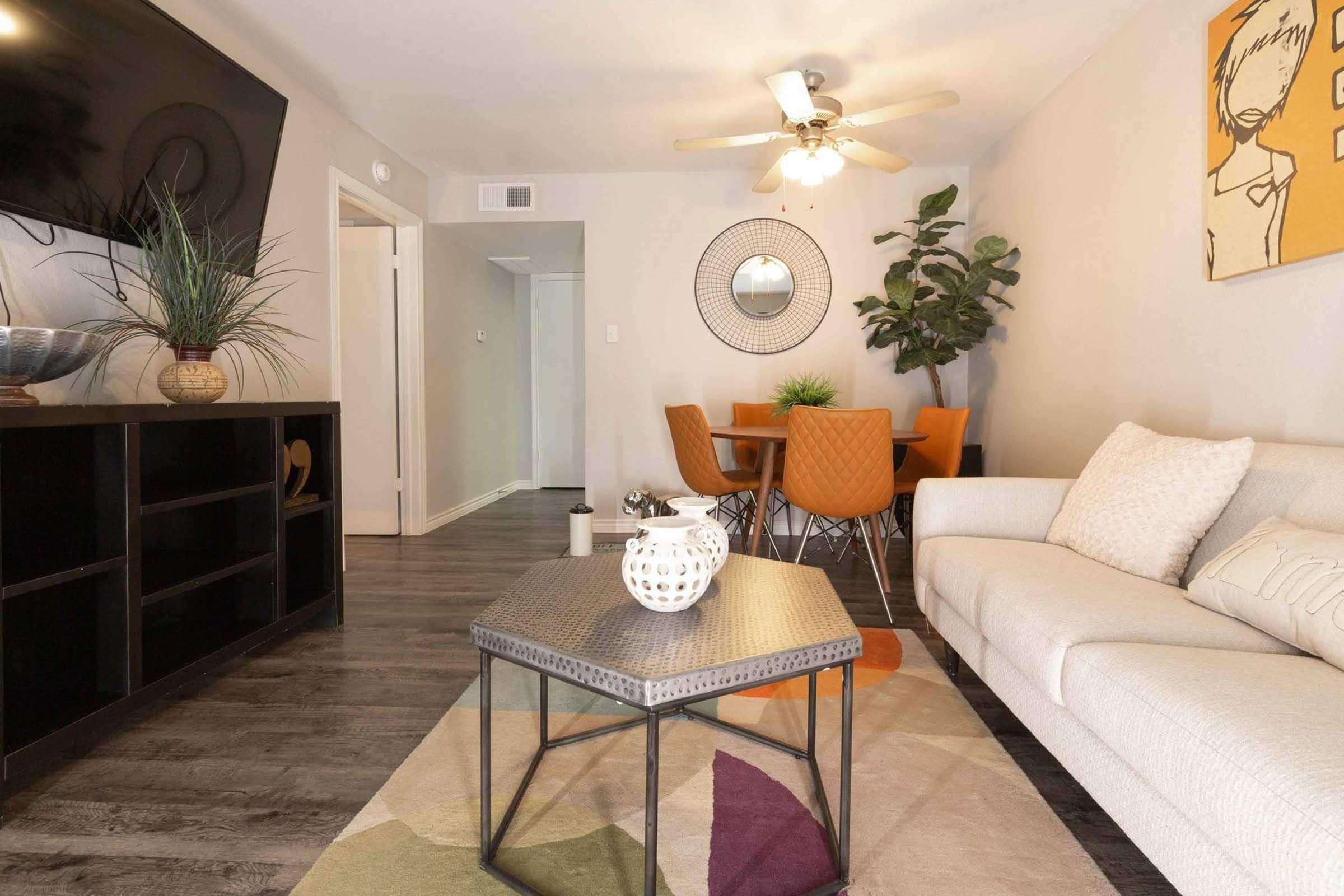 A modern living room featuring a light-colored sofa, a geometric coffee table, and a round mirror on the wall. A round dining table with orange chairs is visible in the background, along with a leafy plant and a decorative bowl on the coffee table. The flooring is dark wood, and there is a ceiling fan above.