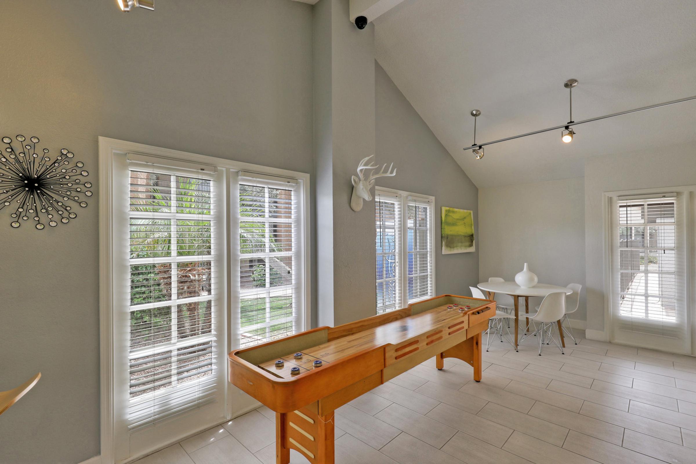 A spacious interior featuring a shuffleboard table, large windows with blinds allowing natural light, modern light fixtures, and a small dining area with white chairs and a table.