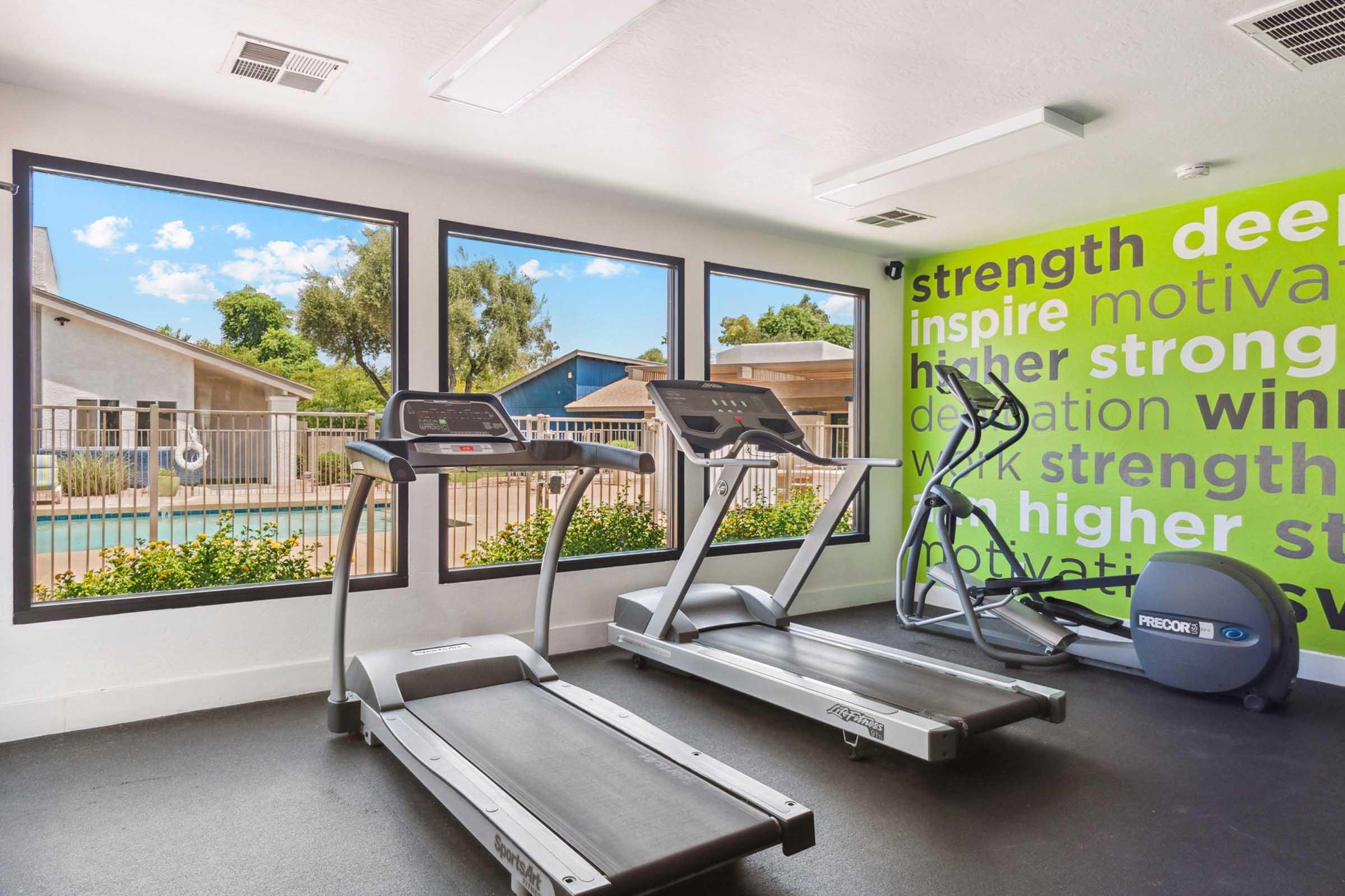 A modern fitness room featuring two treadmills and an elliptical machine, with large windows offering views of an outdoor pool area.