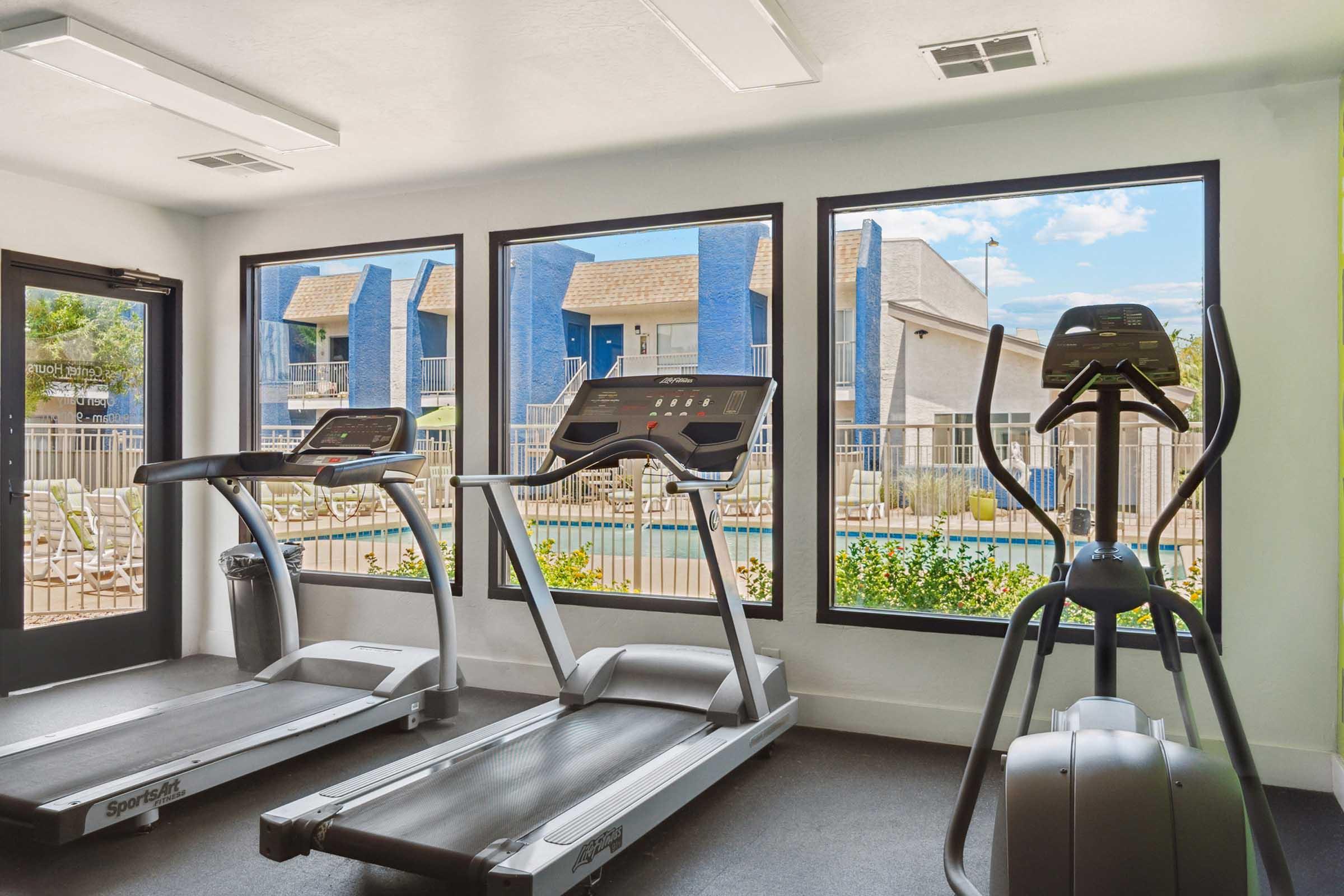 A bright, modern gym with two treadmills and an elliptical machine. Large windows provide a view of a pool area and exterior buildings.