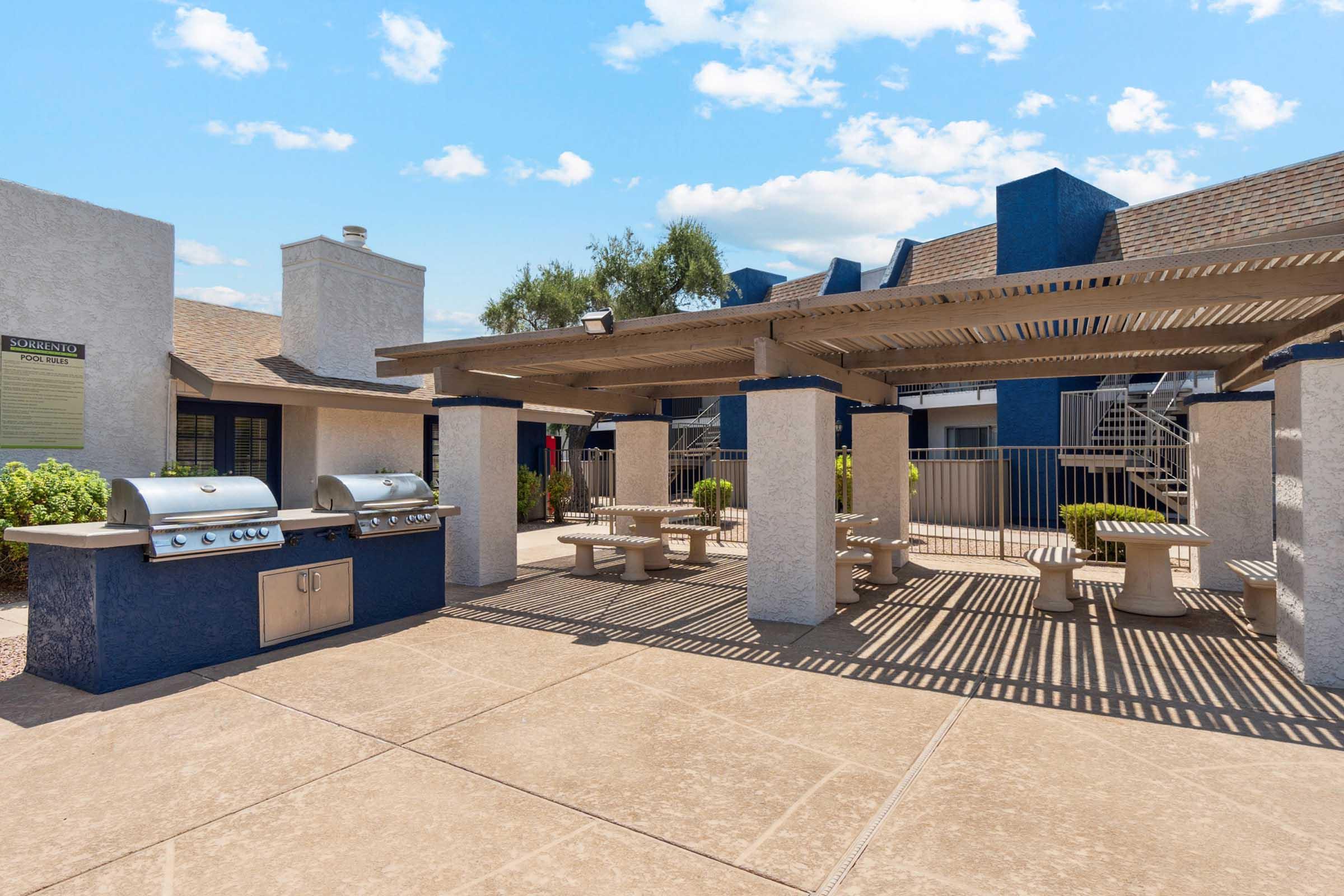 Outdoor patio area featuring two stainless steel grills, picnic tables, and shaded seating under a pergola.