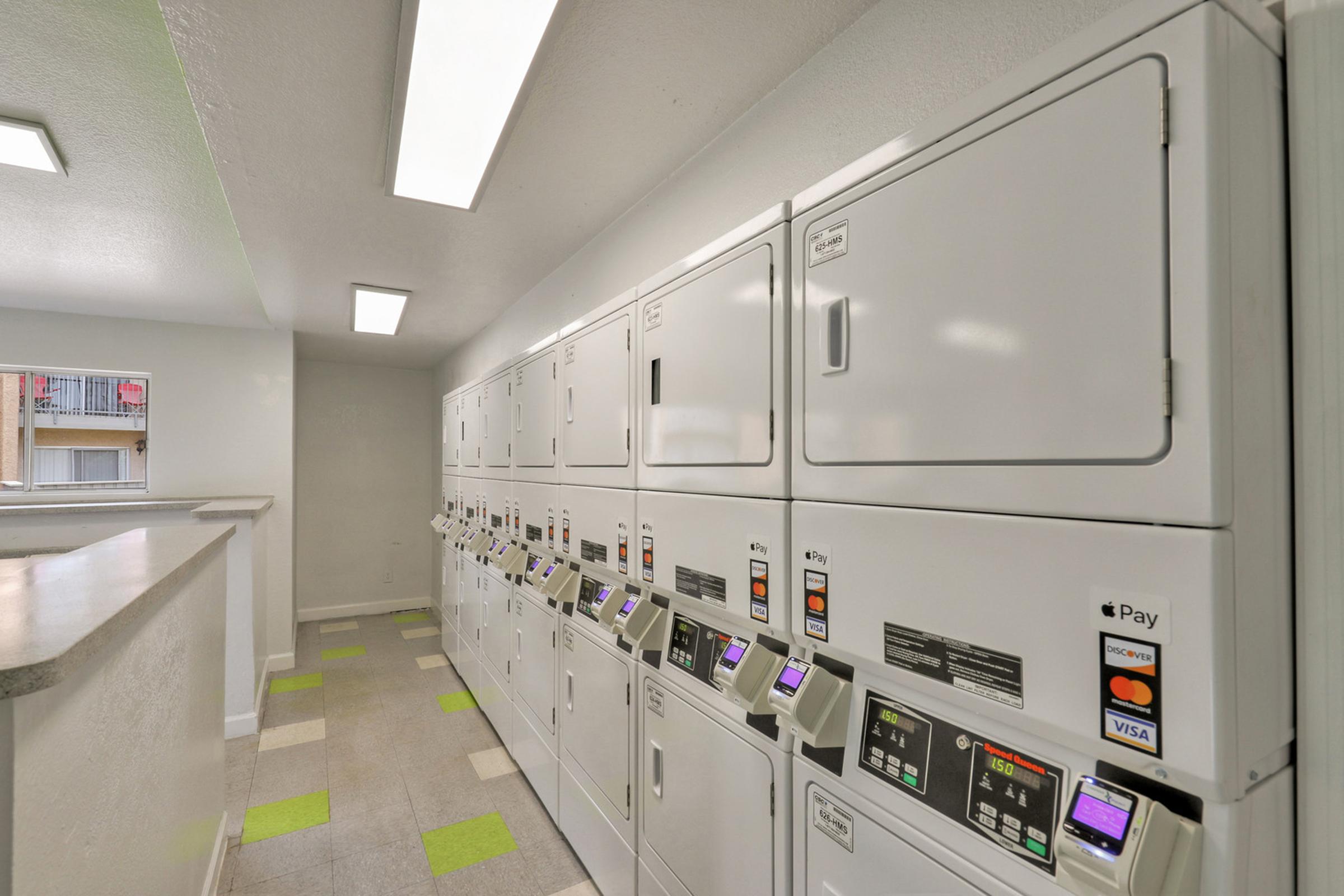 A clean, modern laundry room featuring a row of white coin-operated washing machines and dryers.