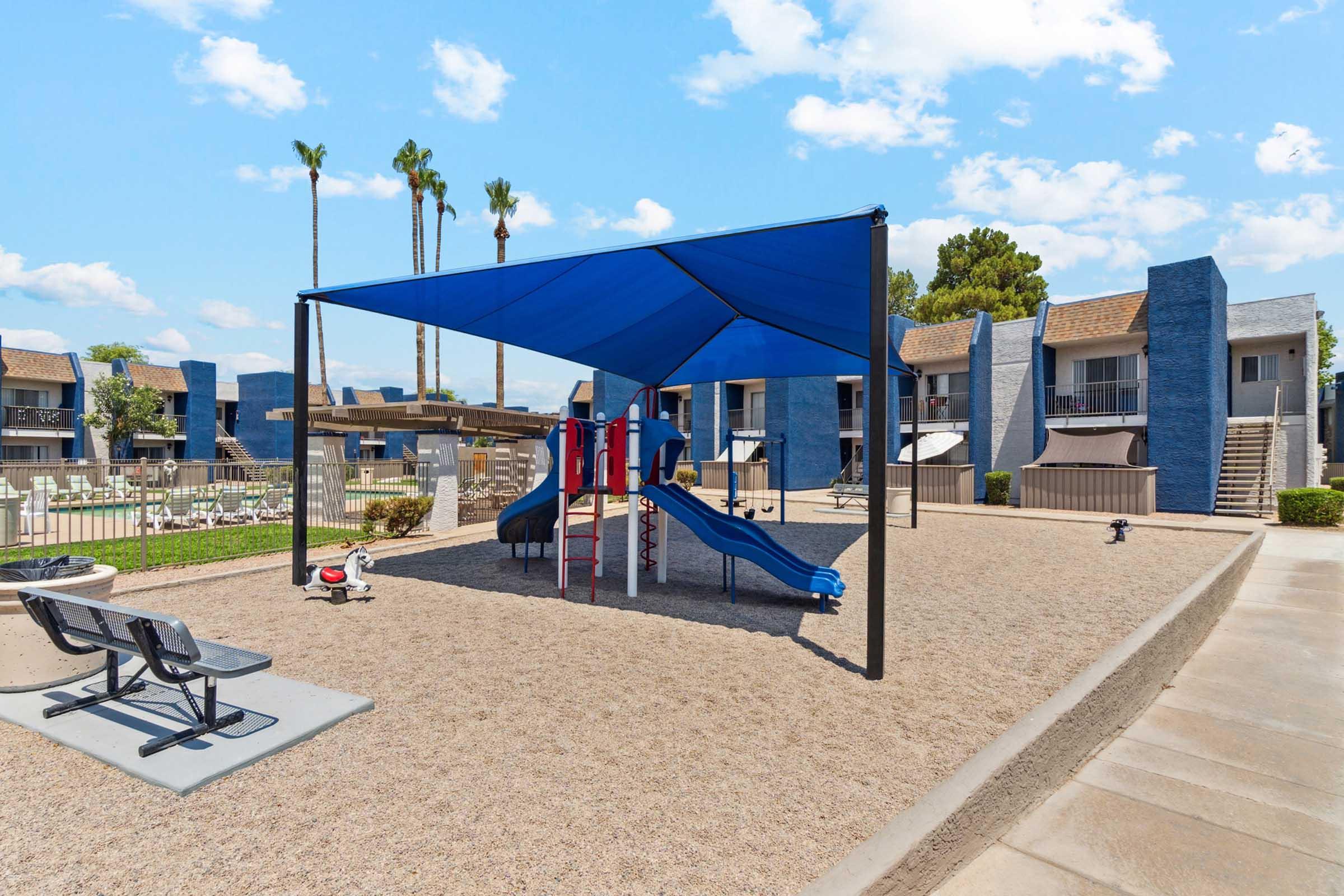 A playground with blue slides and climbing structures is shaded by a large blue canopy.