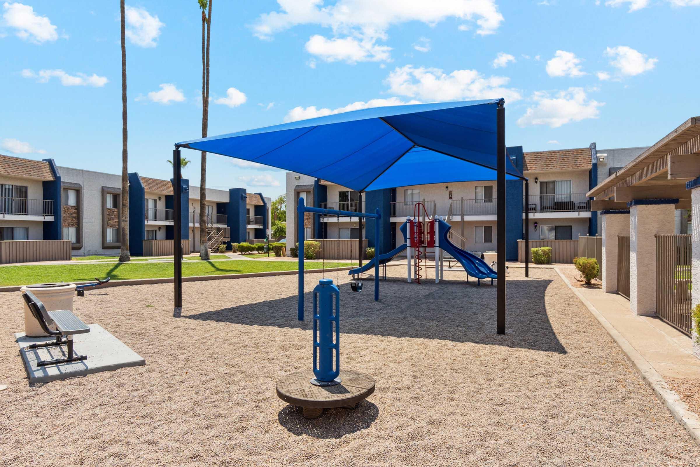A playground area featuring a blue shade structure, slide, and climbing equipment set in a gravel surface.