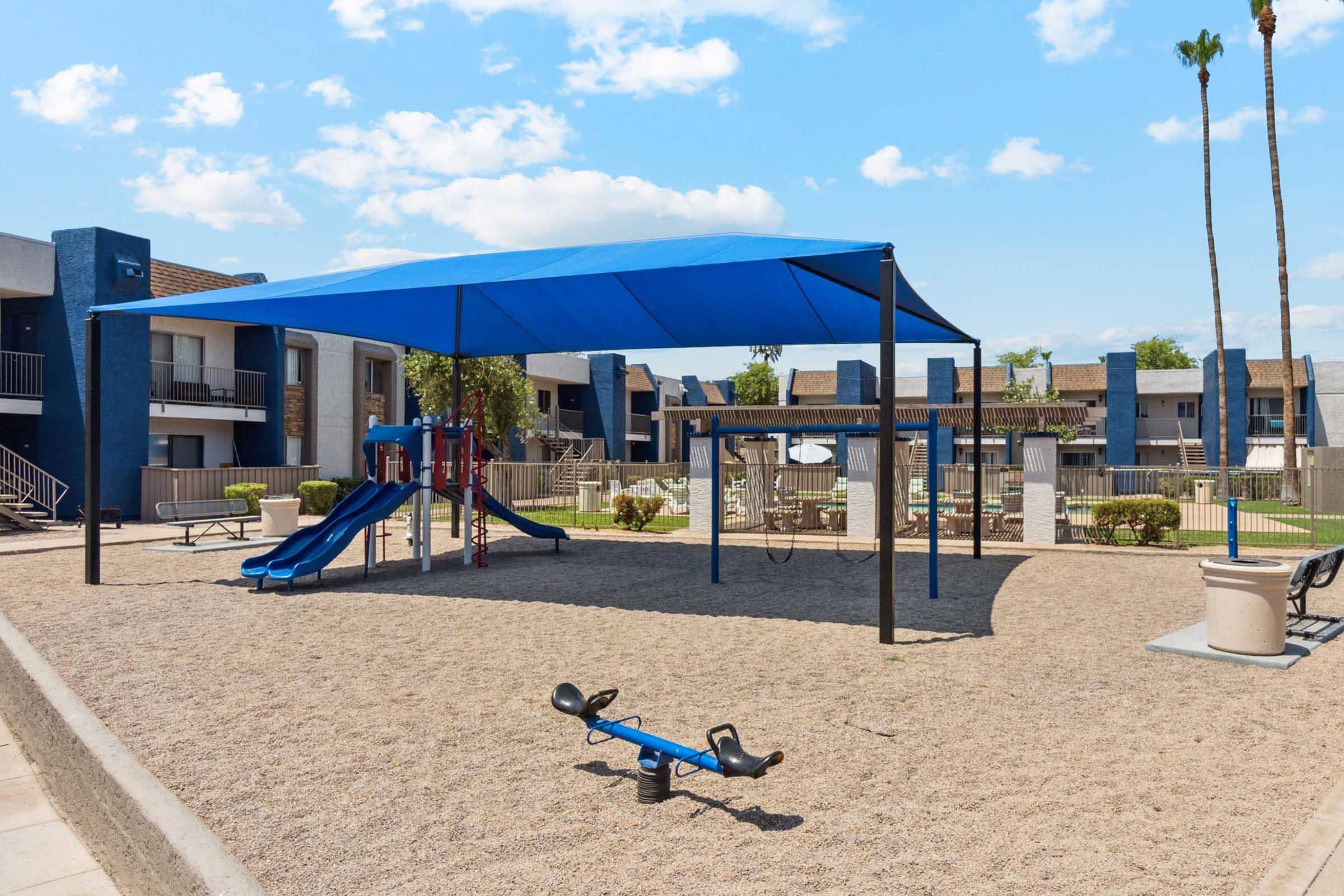 A playground featuring a blue slide, swings, and a seesaw, located in a sandy area.