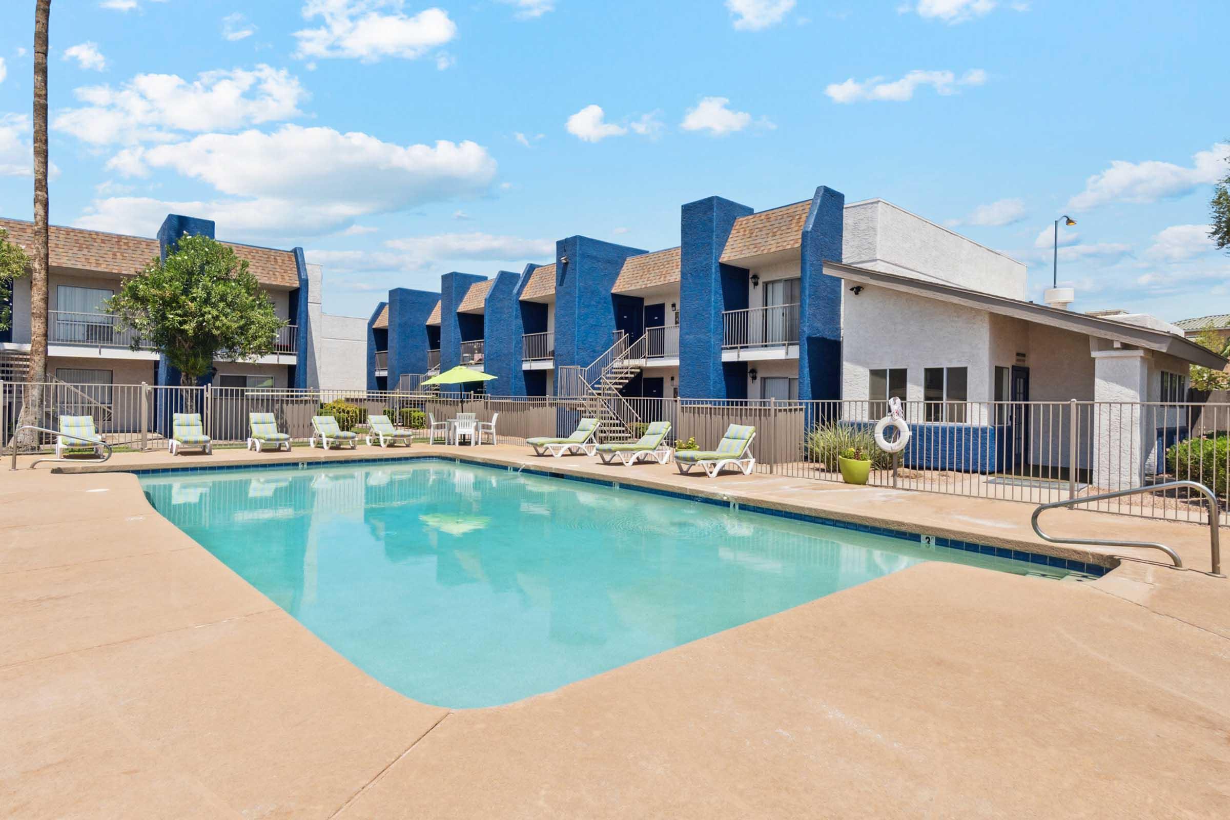 A clear swimming pool surrounded by lounge chairs, with a backdrop of two-story buildings featuring blue and white exteriors.