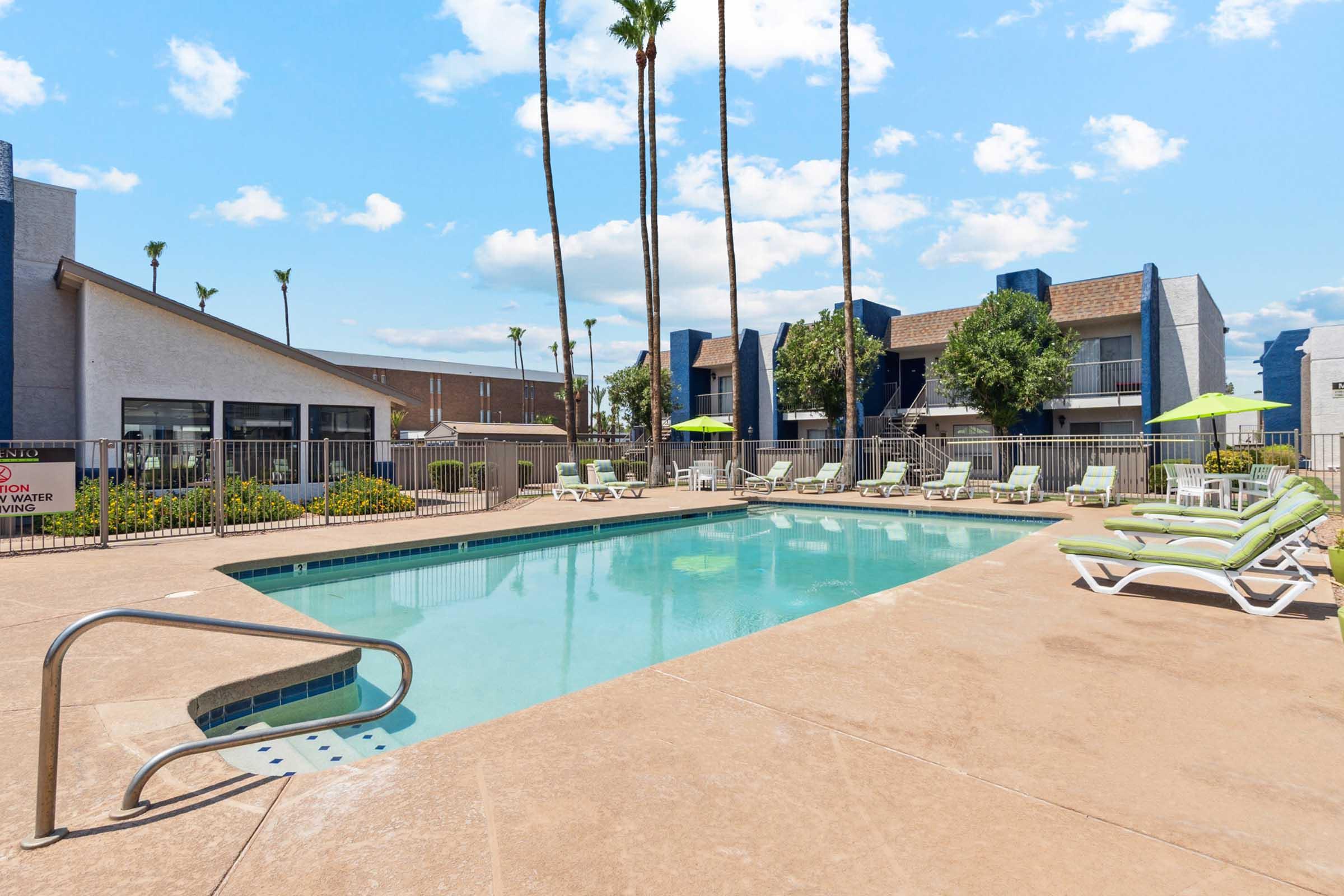 A clear swimming pool surrounded by lounge chairs and palm trees, with a blue sky and fluffy clouds in the background.