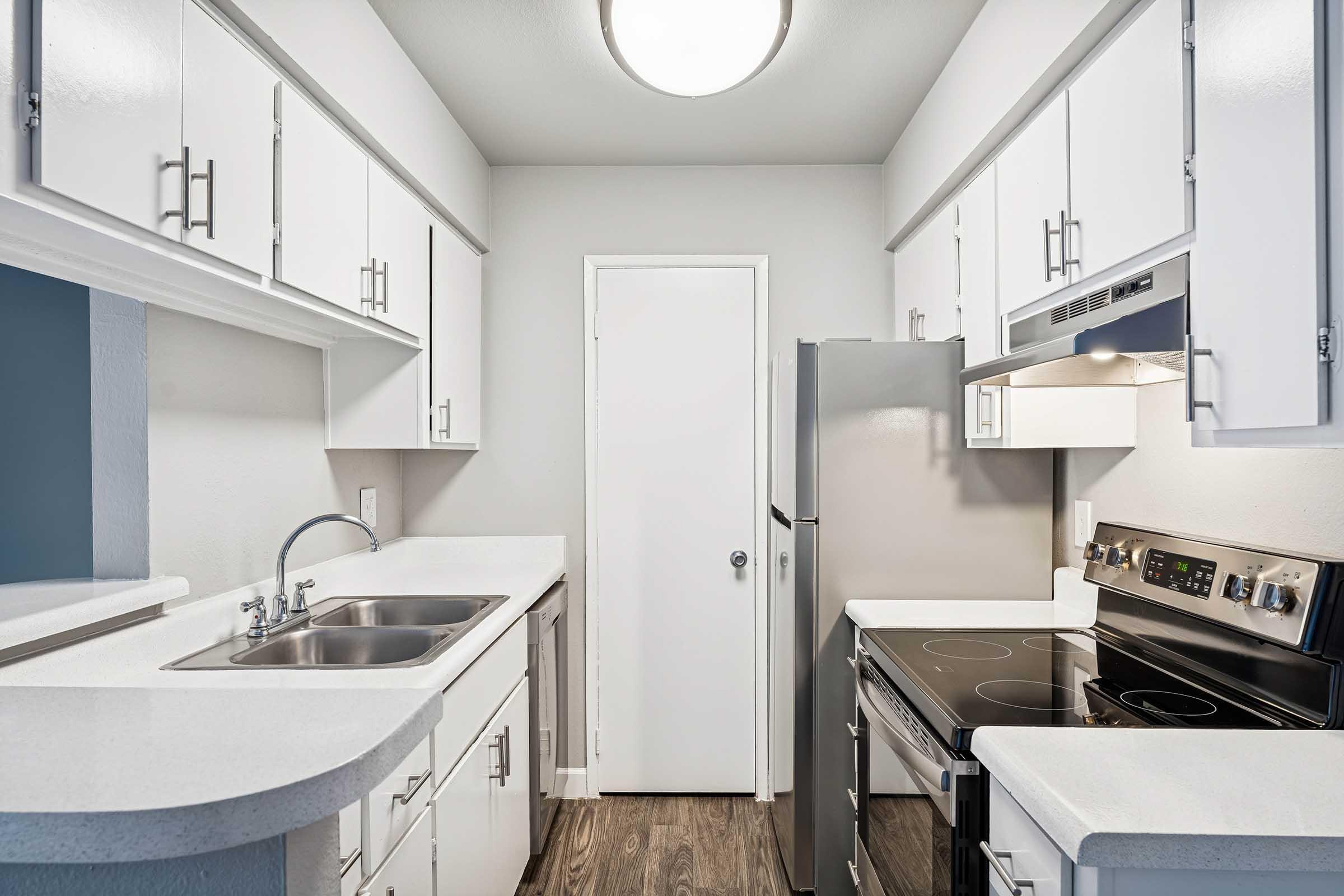 Modern kitchen featuring white cabinetry, a double sink, a stainless steel refrigerator, and an electric stove with an over-the-range microwave. The countertops are light-colored, creating a bright and airy feel. A door leads to an adjacent room, and the space has wood-like flooring.