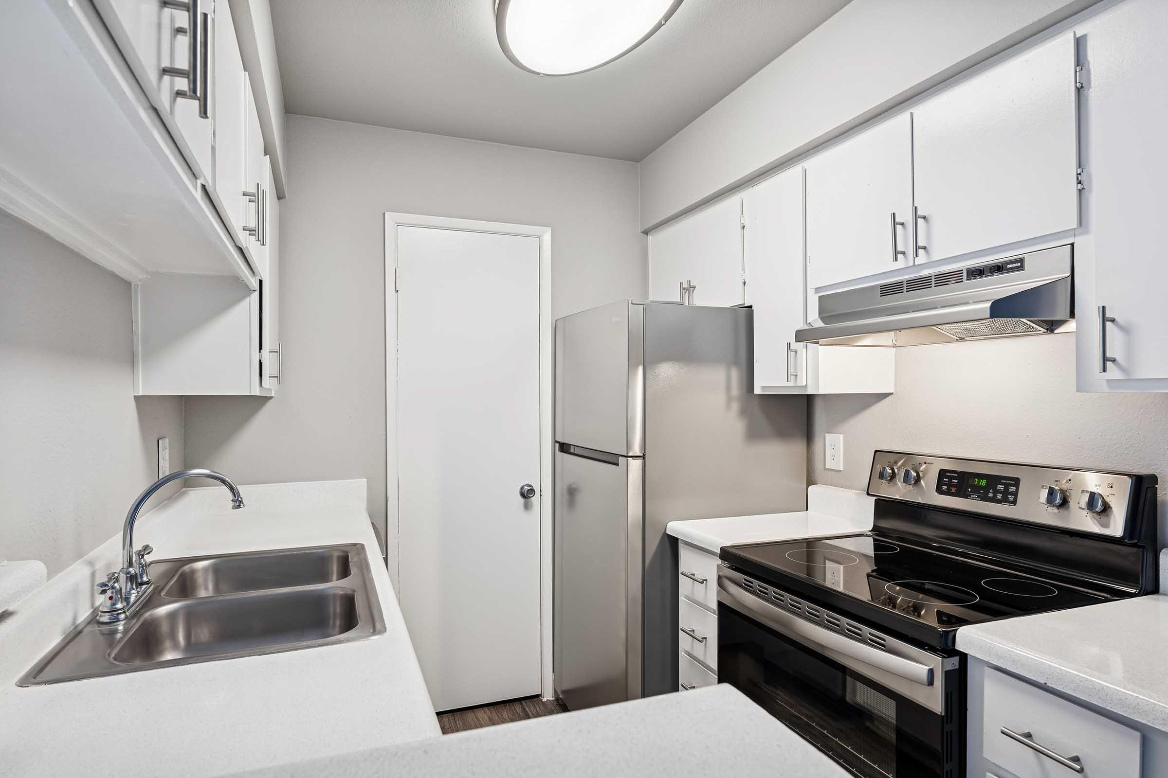 A modern kitchen featuring a stainless steel refrigerator, stove, and a double sink. The cabinetry is white with simple hardware, and the walls are painted a light color. A door leads to another room, and there is an overhead light fixture providing illumination. The countertops are light-colored and clean.