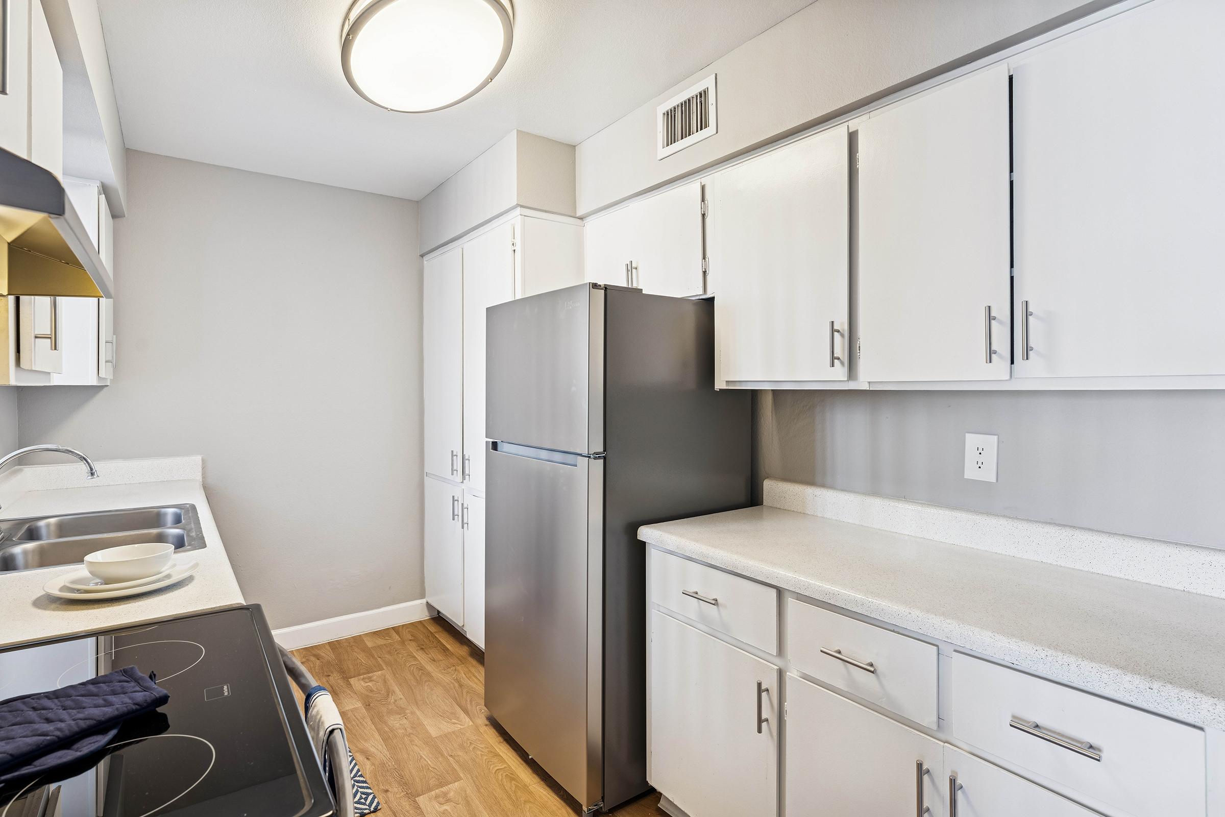 Modern kitchen featuring white cabinets and a stainless steel refrigerator. The countertop is light-colored with a sink and stovetop visible. The space has a clean, organized look with a light fixture above.