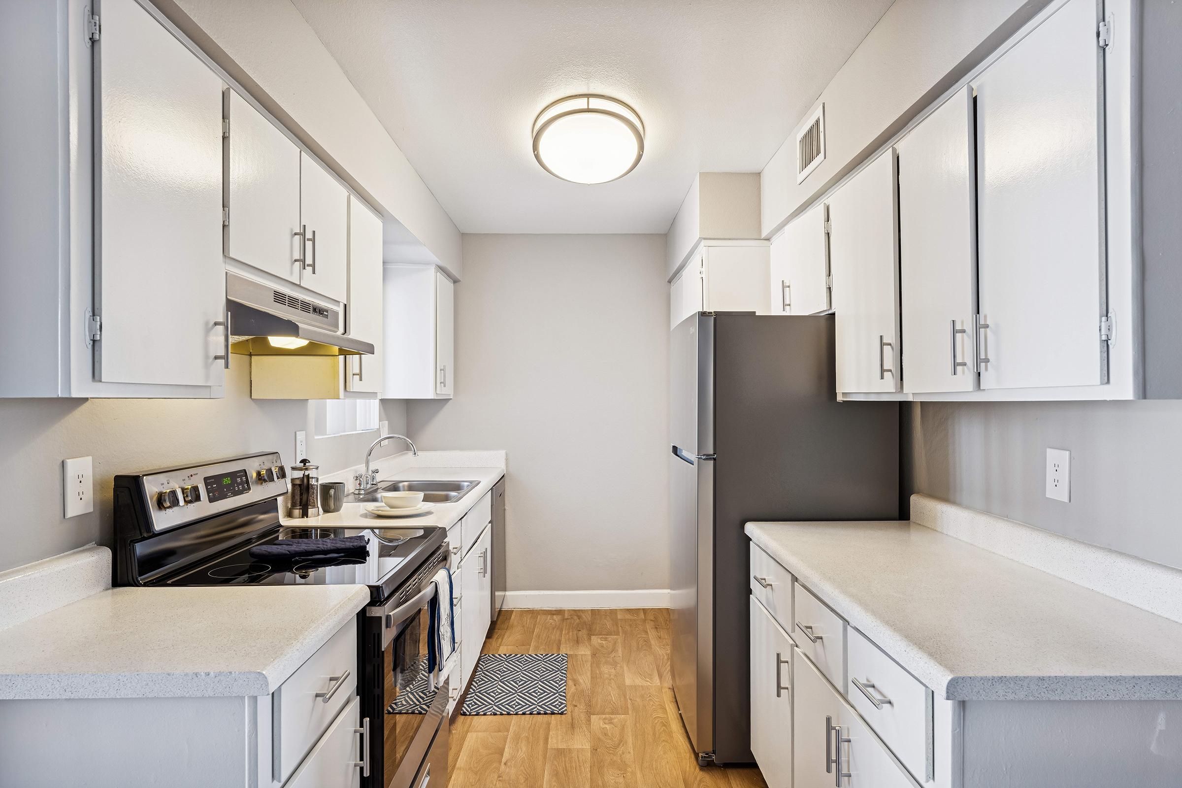 Modern kitchen featuring white cabinetry, stainless steel appliances, and a gray refrigerator. The space is well-lit with a ceiling light, and includes an oven, stovetop, and sink. Light wood flooring complements the bright, clean design. Countertops are clear with a small rug by the sink.