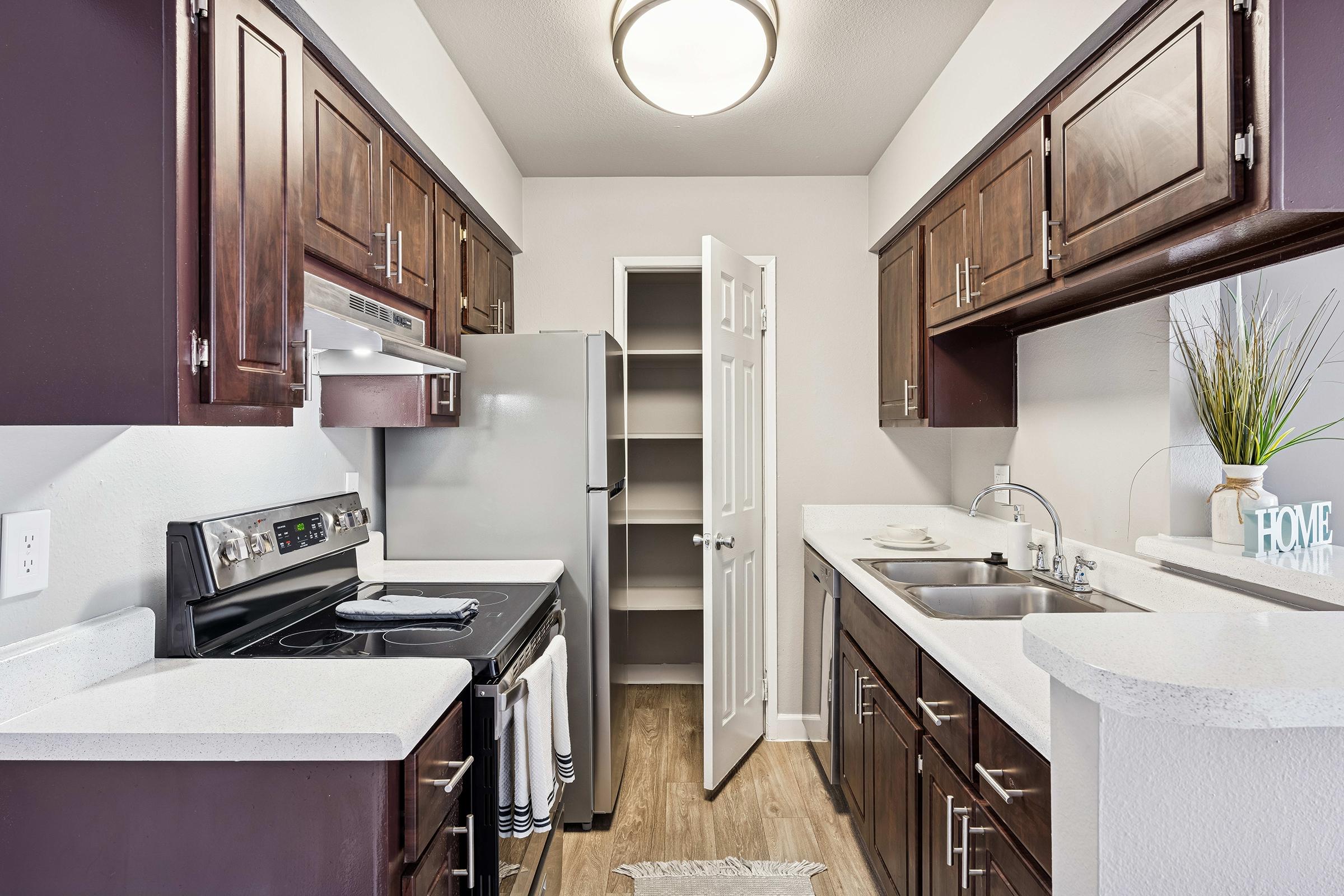 A modern kitchen featuring dark wood cabinetry, stainless steel appliances, and a white countertop. It includes an oven, stovetop, and double sink, with an open pantry door visible. The space is well-lit, showcasing a comfortable and functional layout.