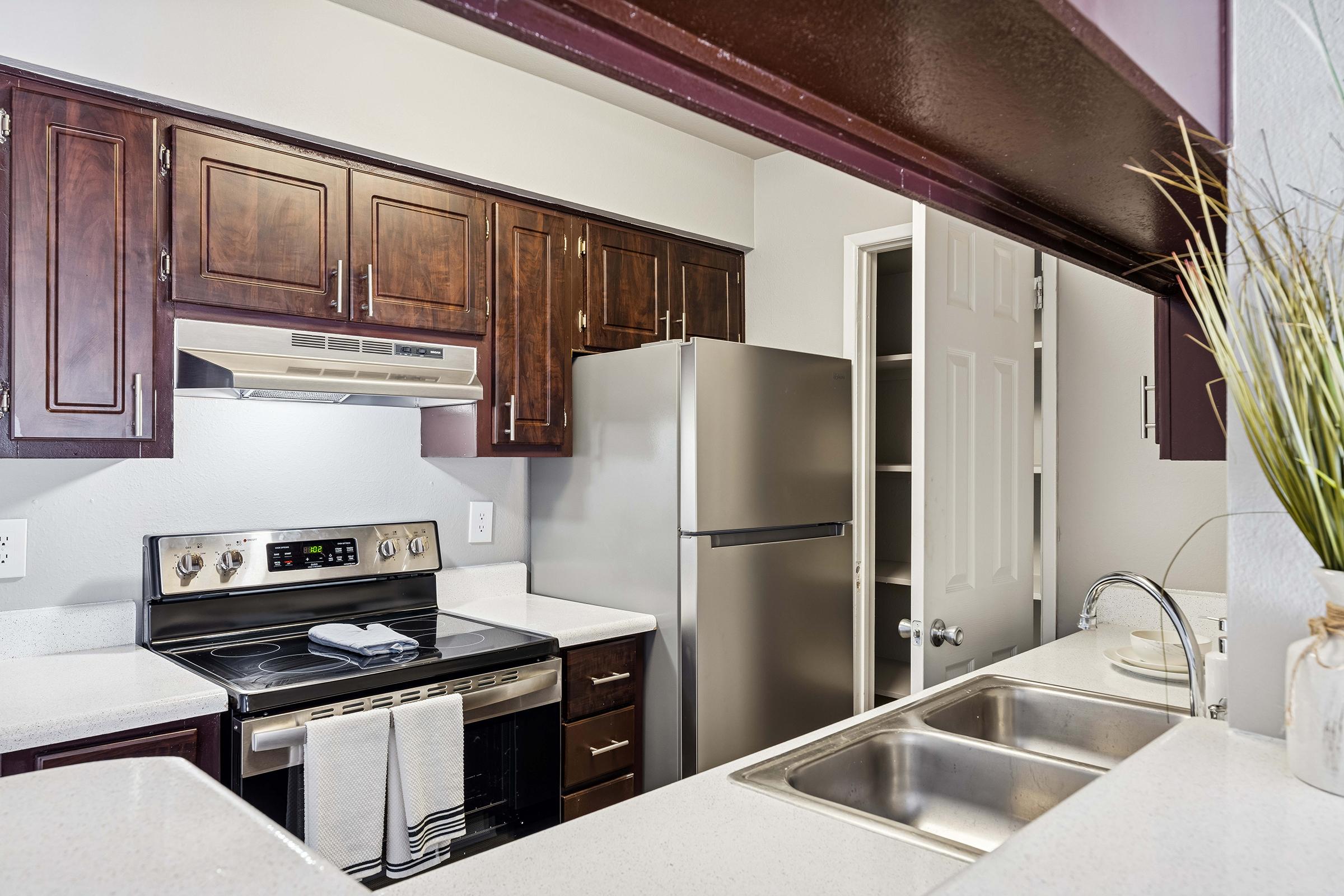 A modern kitchen featuring dark wooden cabinets, stainless steel appliances including a stove and refrigerator, white countertops, and a dual sink. The design includes a small opening to another room, adding an open feel to the space. A plant is displayed on the counter for decoration.