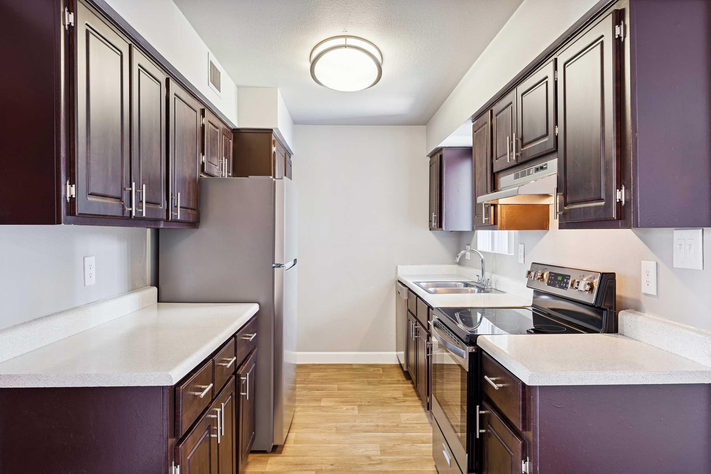 A modern kitchen featuring dark wood cabinets, stainless steel appliances including a refrigerator and stove, a light-colored countertop, and wood-look flooring. The space is well-lit with a ceiling fixture, and the layout includes an open area with ample counter space and a sink near the stove.