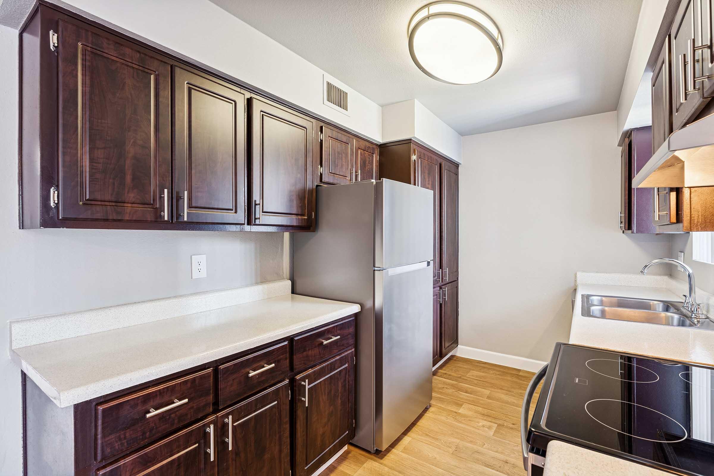 Modern kitchen featuring dark wooden cabinets, a stainless steel refrigerator and stove, and a light countertop. The space is well-lit with natural light and has a neutral-colored wall. The flooring is a light wood finish, adding warmth to the overall design.