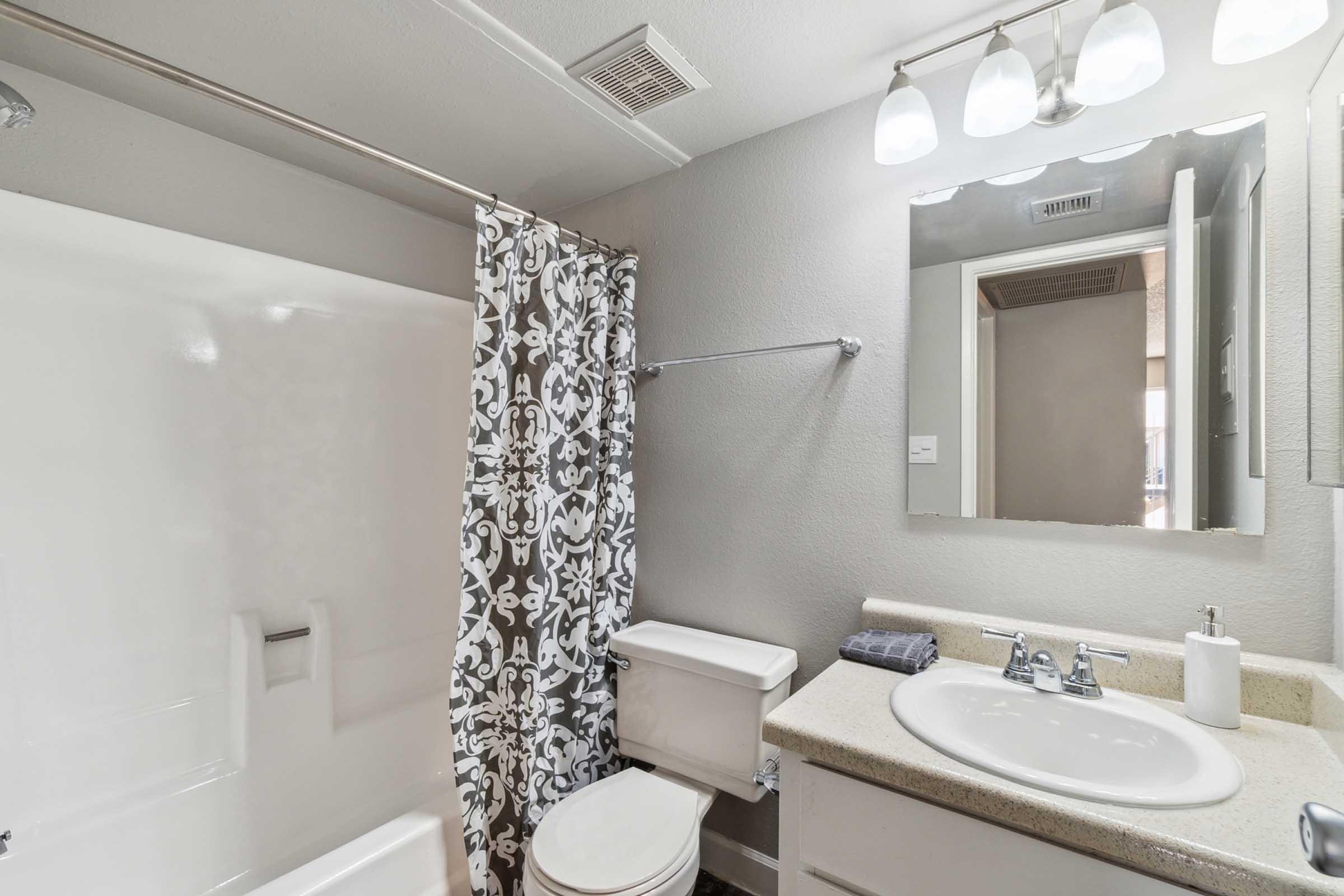 A modern bathroom featuring a white bathtub with a shower, a decorative shower curtain in black and white, a countertop with a sink, a mirror above the sink, and lighting fixtures.
