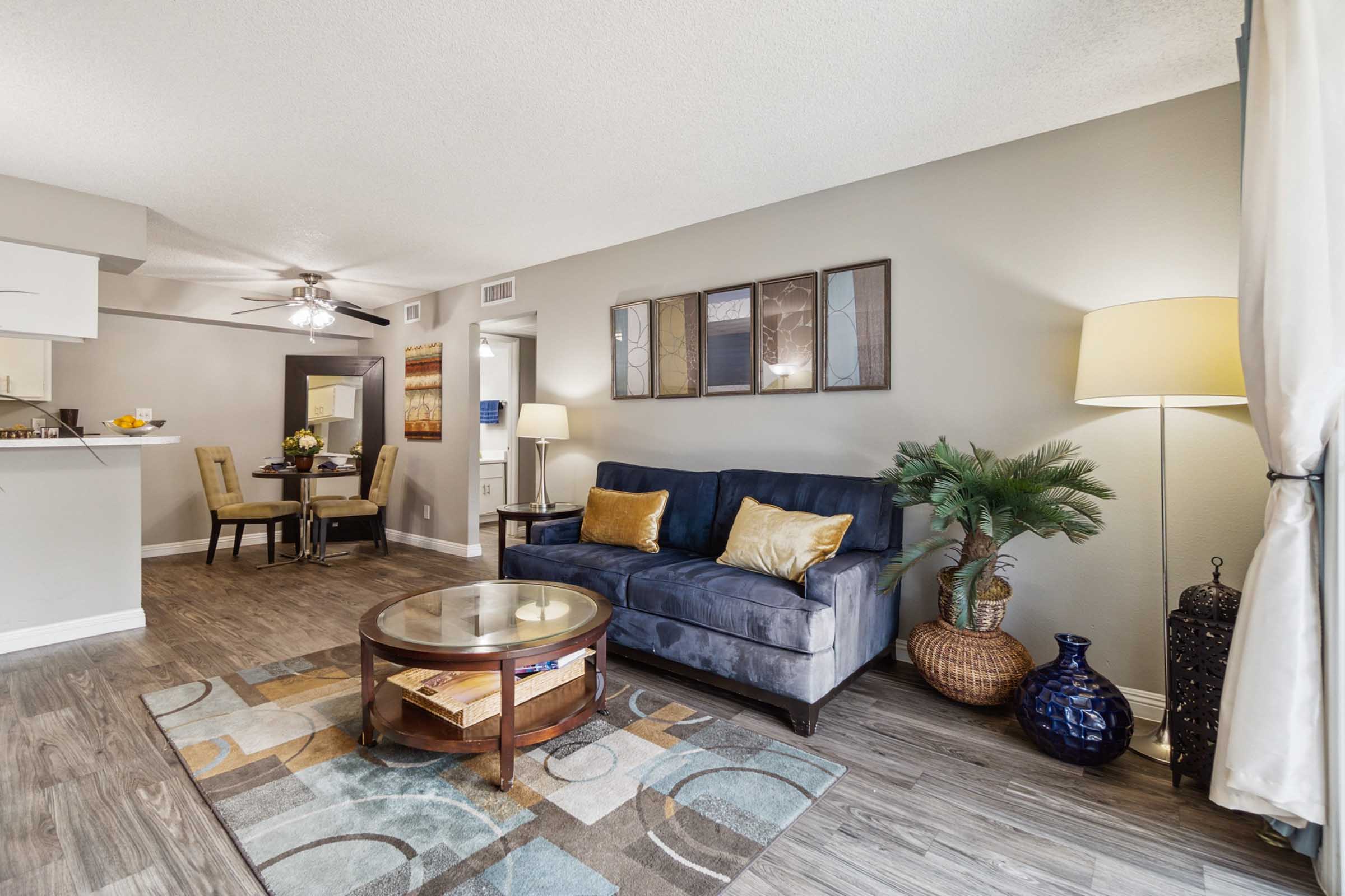 A cozy living room featuring a blue sofa with decorative cushions, a round coffee table, and a tall lamp. There is a potted plant beside the sofa and artwork on the wall. The space has wood-style flooring and a dining area visible in the background with a small table and chairs.