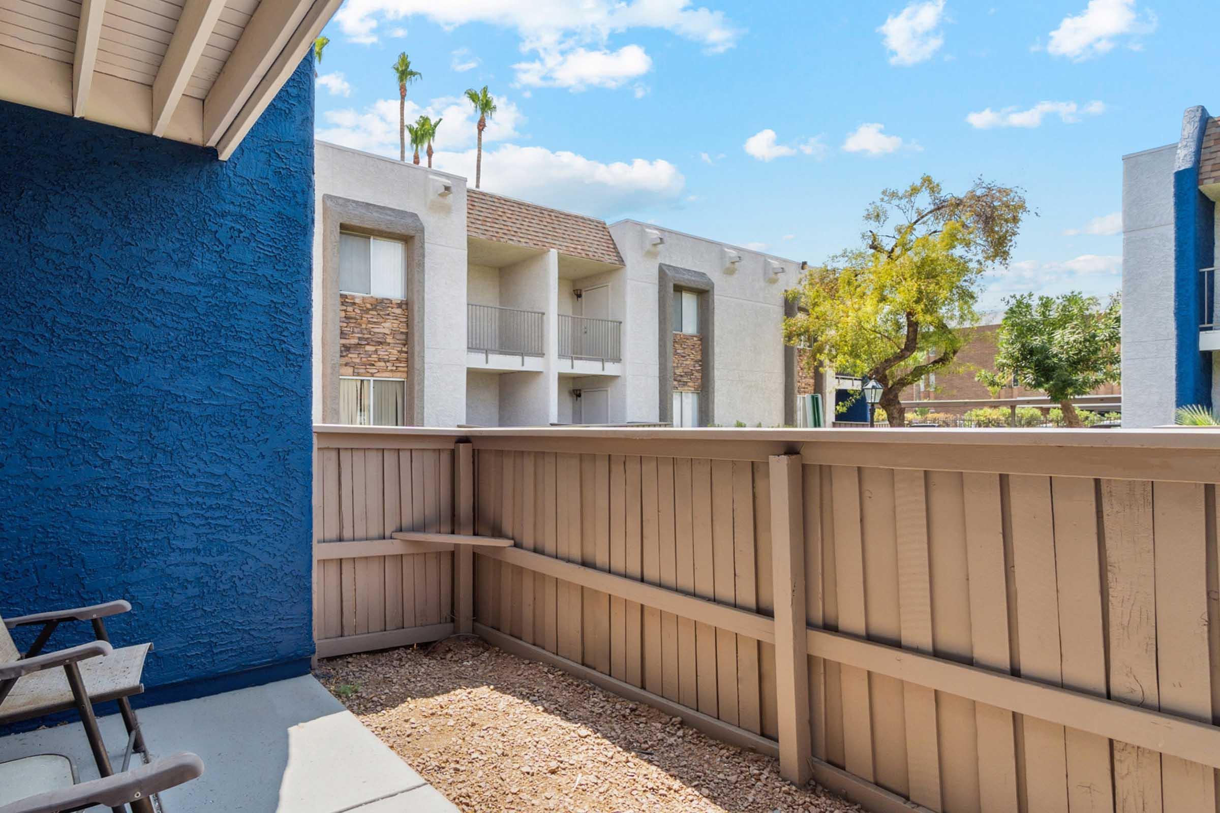 View from a patio featuring a wooden fence and seating area, with a glimpse of a nearby apartment complex and palm trees under a partly cloudy sky