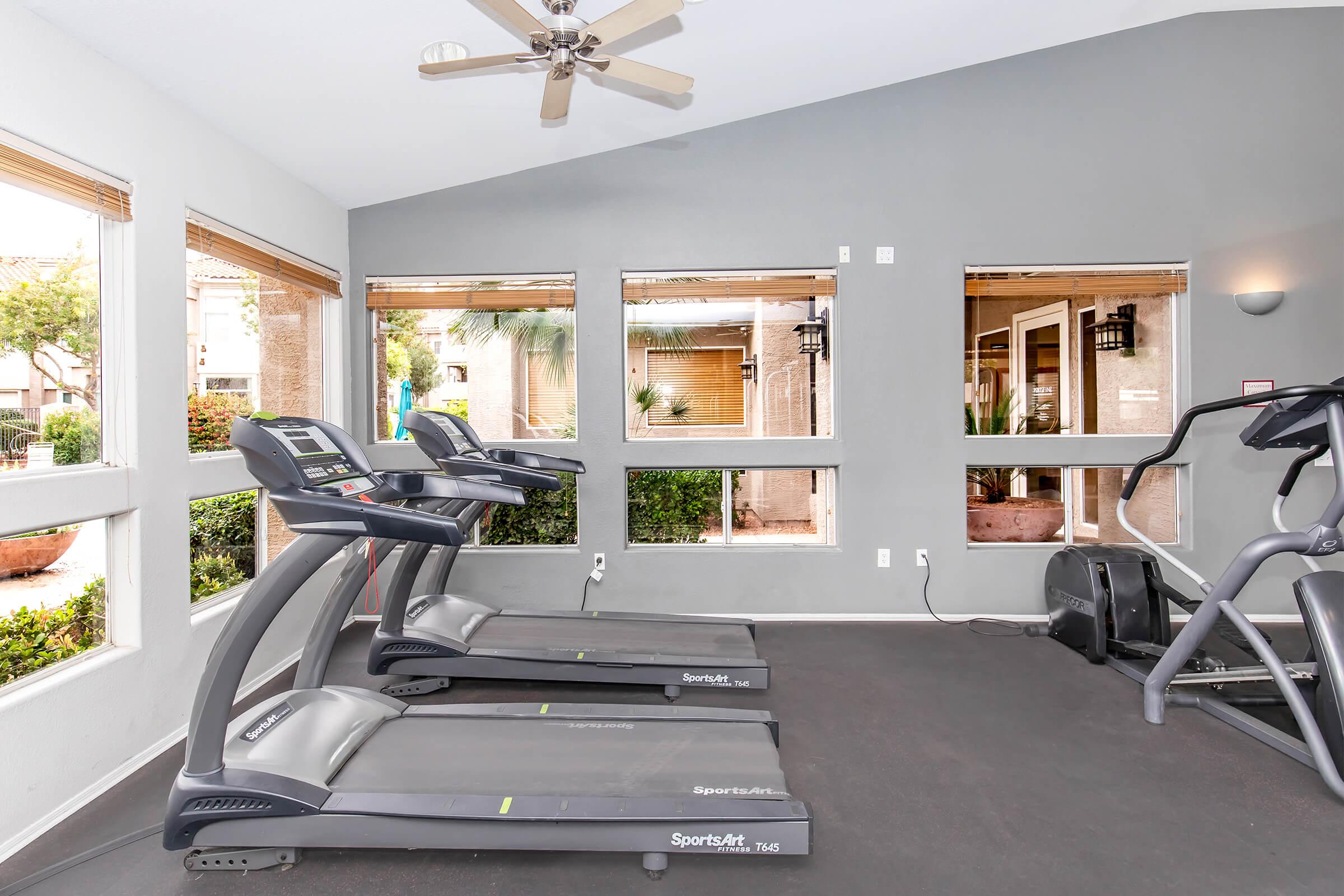 A fitness room featuring three treadmills and an elliptical machine, with large windows allowing natural light. The walls are painted gray, and a ceiling fan is visible. The room has a clean and modern design, with greenery visible outside the windows, creating a pleasant workout atmosphere.