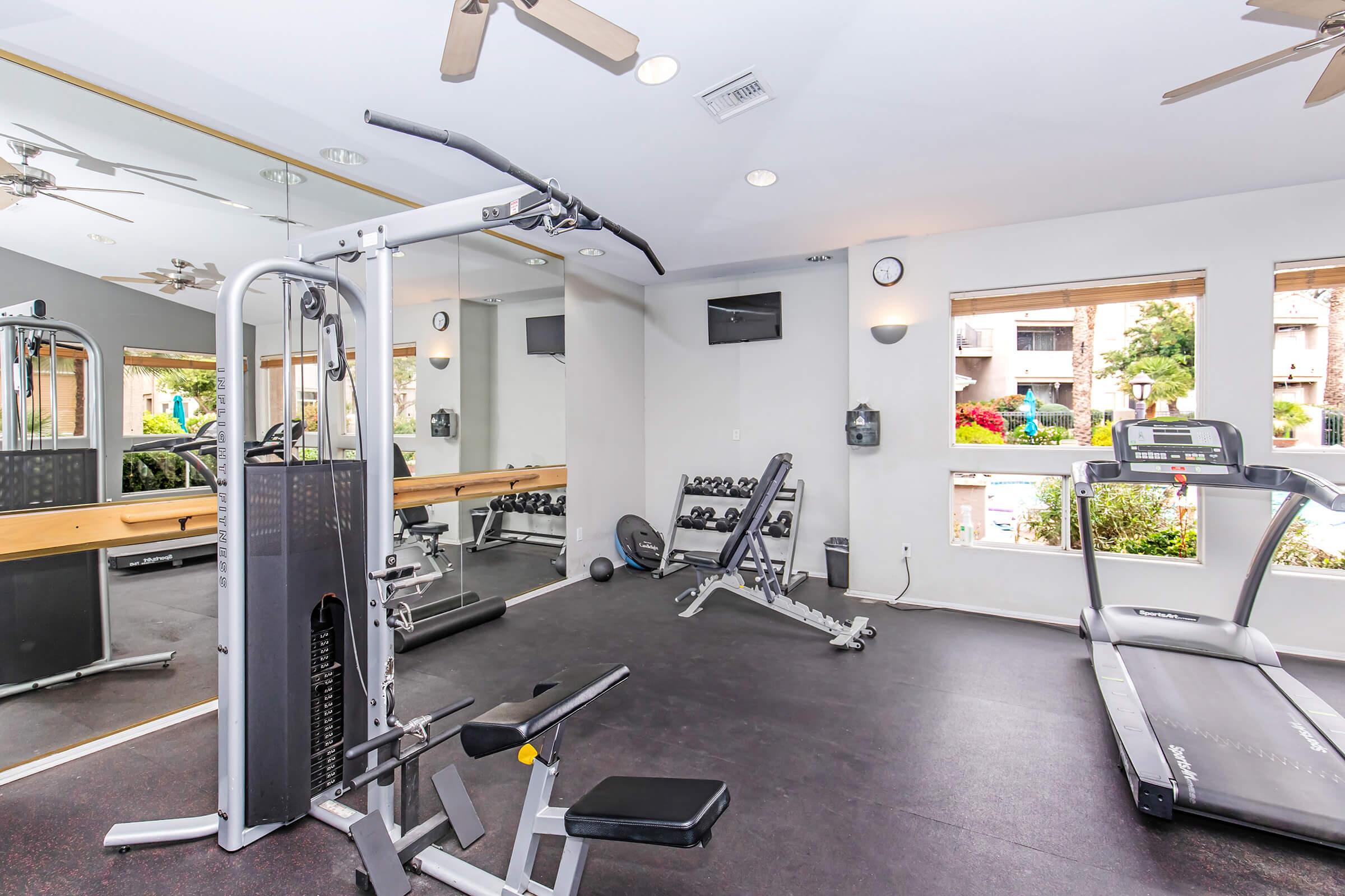 A well-equipped gym area featuring a treadmill, weightlifting machine, free weights, and large mirrors. The bright space has natural light coming through windows, with fitness equipment arranged neatly on a dark floor.