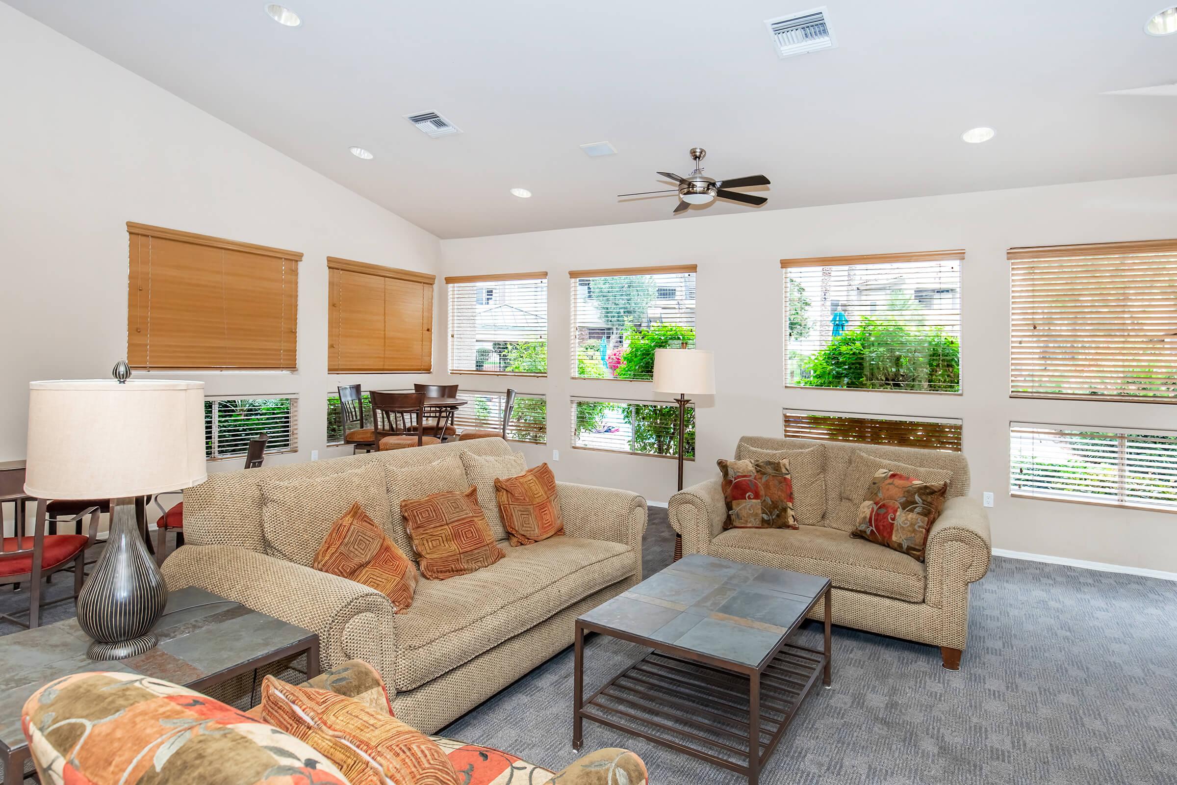 A cozy living area featuring a light-colored couch with patterned cushions, a coffee table, and a side table with a lamp. There are large windows with blinds allowing natural light, and a dining area visible in the background with a wooden table and chairs. A ceiling fan adds to the comfort of the space.