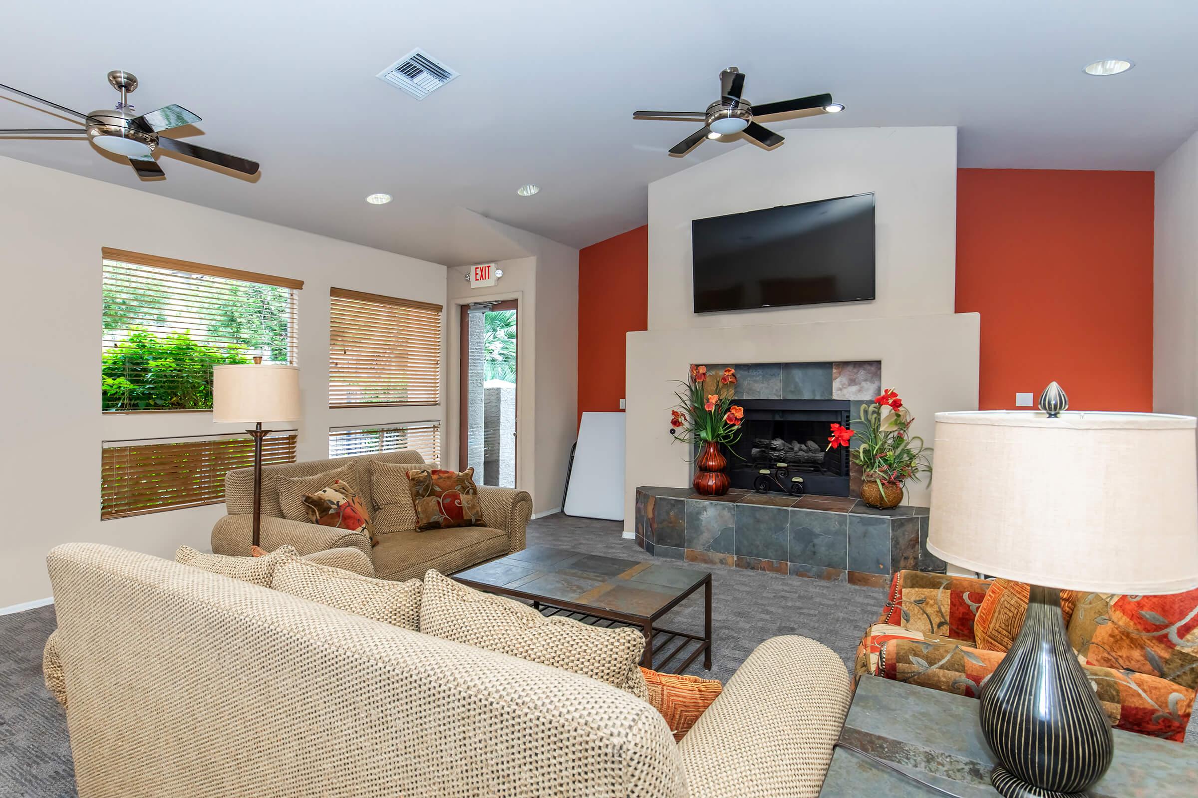A cozy living room featuring a comfortable seating arrangement with beige and patterned sofas, a stone fireplace, and a mounted flat-screen TV. Natural light filters through window blinds, and decorative lamps and plants add a warm touch to the inviting space.