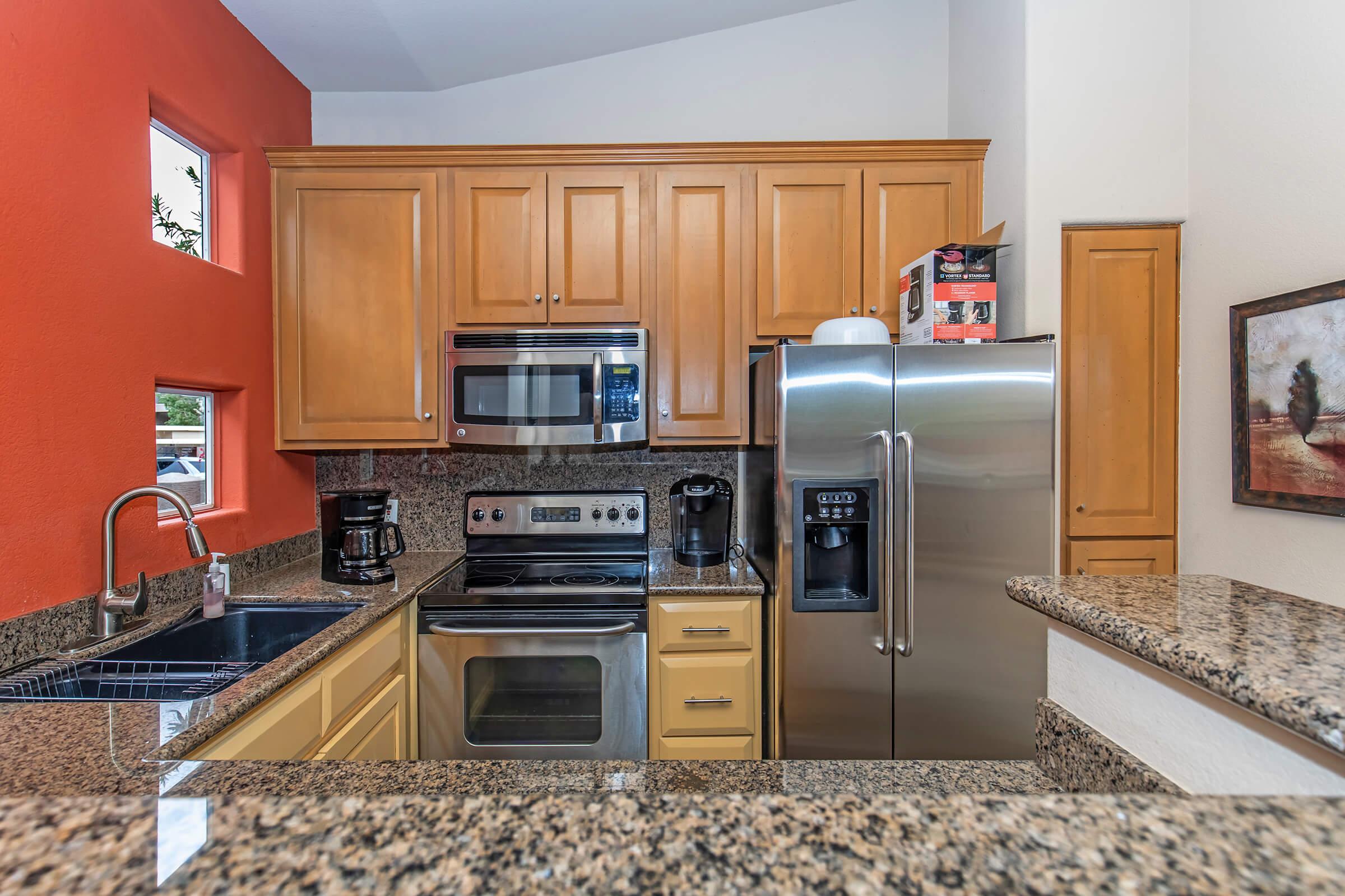 A modern kitchen with wooden cabinetry and granite countertops. It features stainless steel appliances, including a refrigerator, stove, microwave, and coffee maker. A bright red accent wall adds a pop of color, and the area is well-lit with natural light from a window. Decorative artwork is visible on the wall.