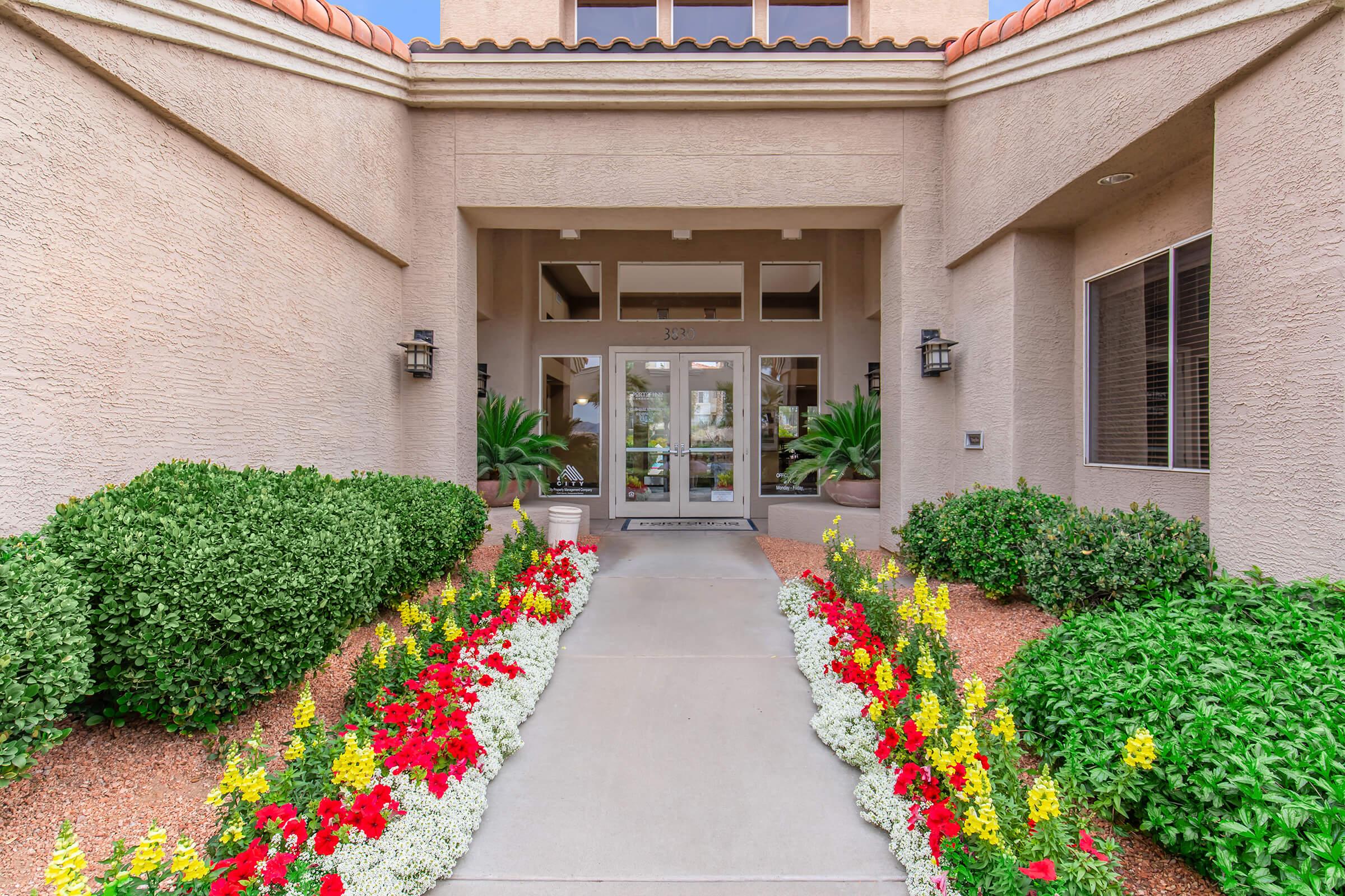 Entrance to a building featuring a pathway lined with colorful flowerbeds, including red, yellow, and white flowers. The facade of the building has a smooth texture with neutral colors and is accented by large windows and potted plants on either side of the entrance.