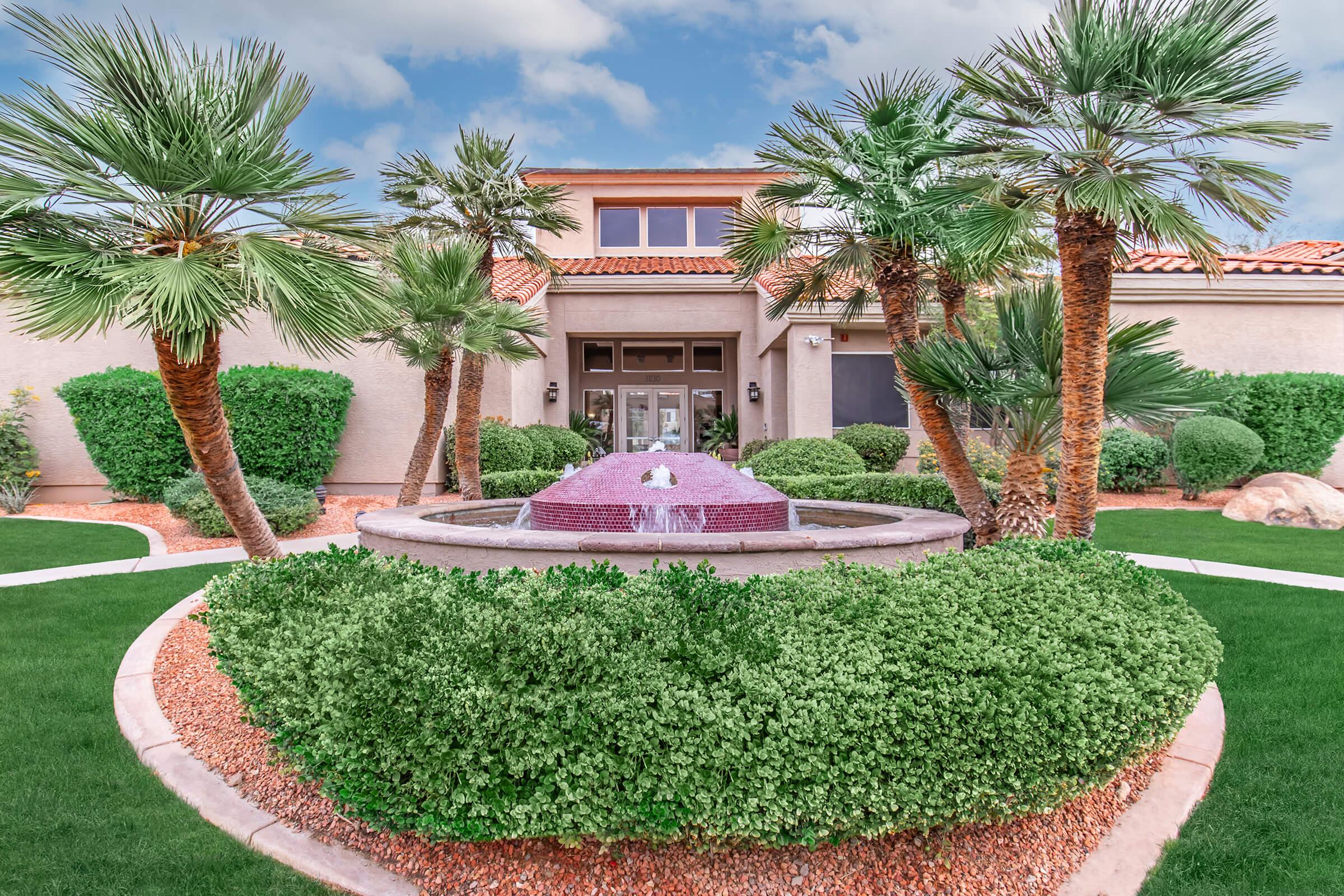 A beautifully landscaped front yard featuring a circular fountain surrounded by lush green hedges and palm trees, with a residential home in the background. The sky is partly cloudy, adding a serene atmosphere to the scene.
