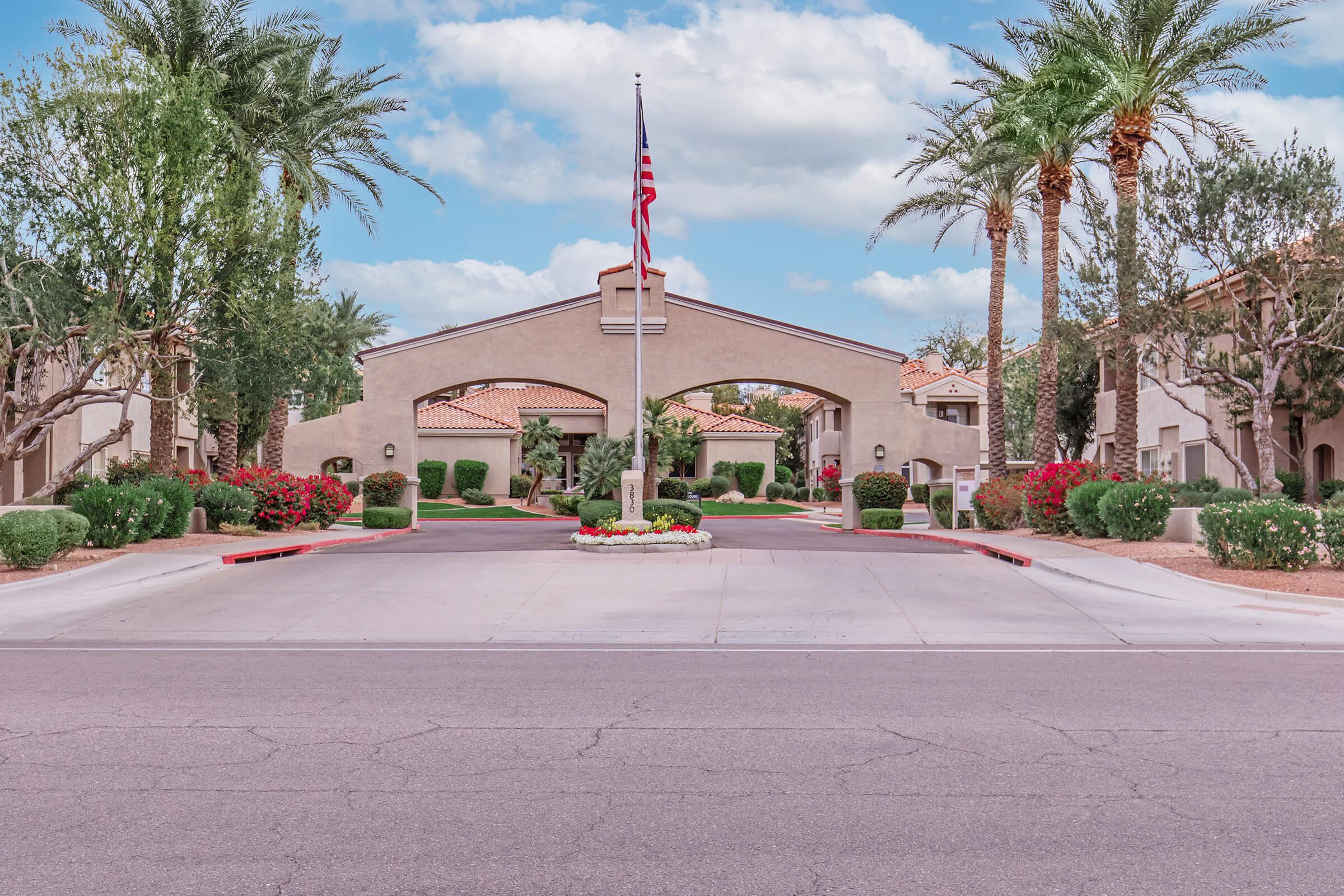 A landscaped entrance to a residential community featuring an arched structure, an American flag on a flagpole, and vibrant flower beds. Palm trees line the pathway, framed by well-maintained greenery. The scene captures a sunny day with a clear blue sky.