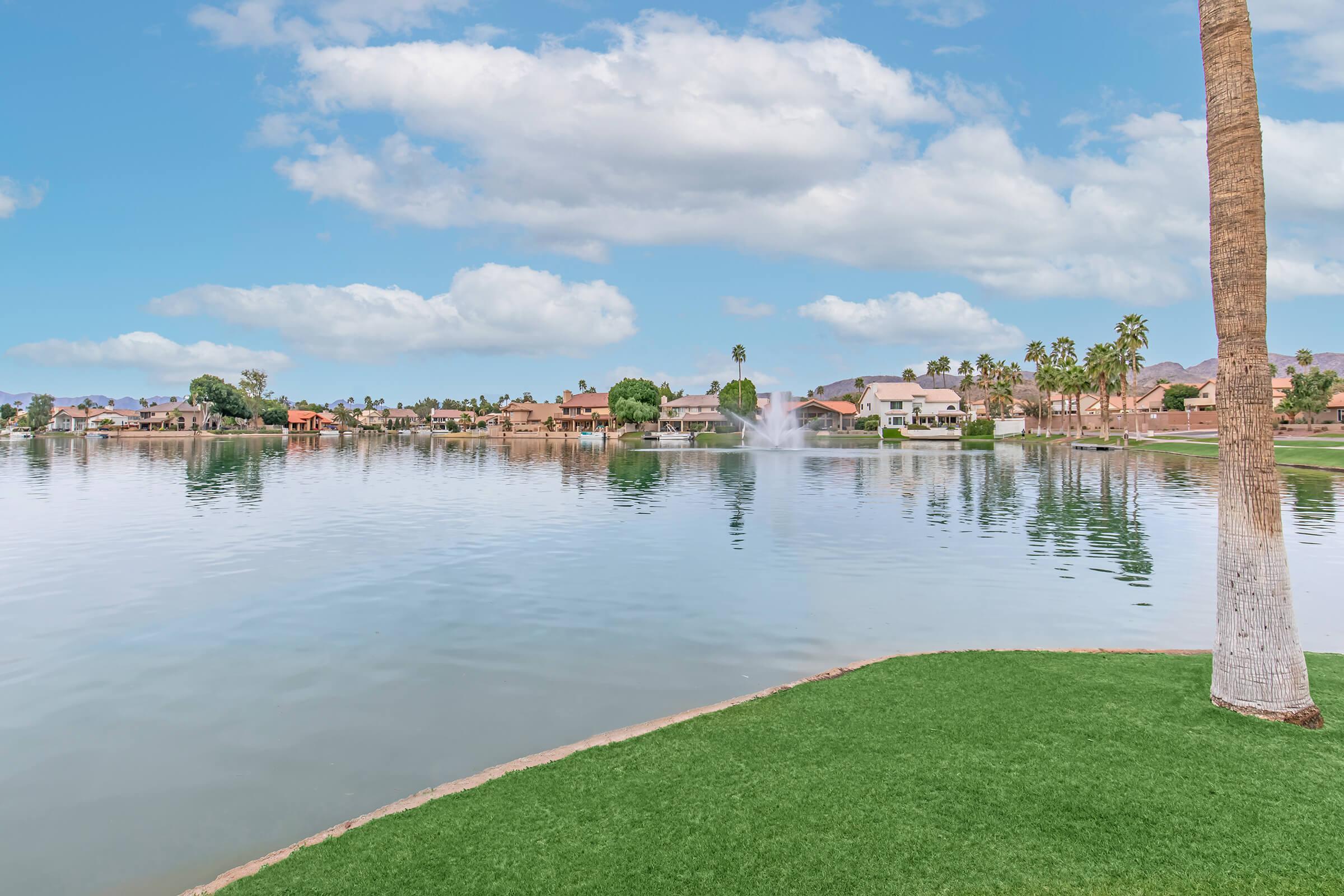 A serene view of a lake surrounded by palm trees and lush green grass. In the background, picturesque homes line the shore, and a fountain creates a gentle spray in the water. The sky is partly cloudy, reflecting a tranquil atmosphere.