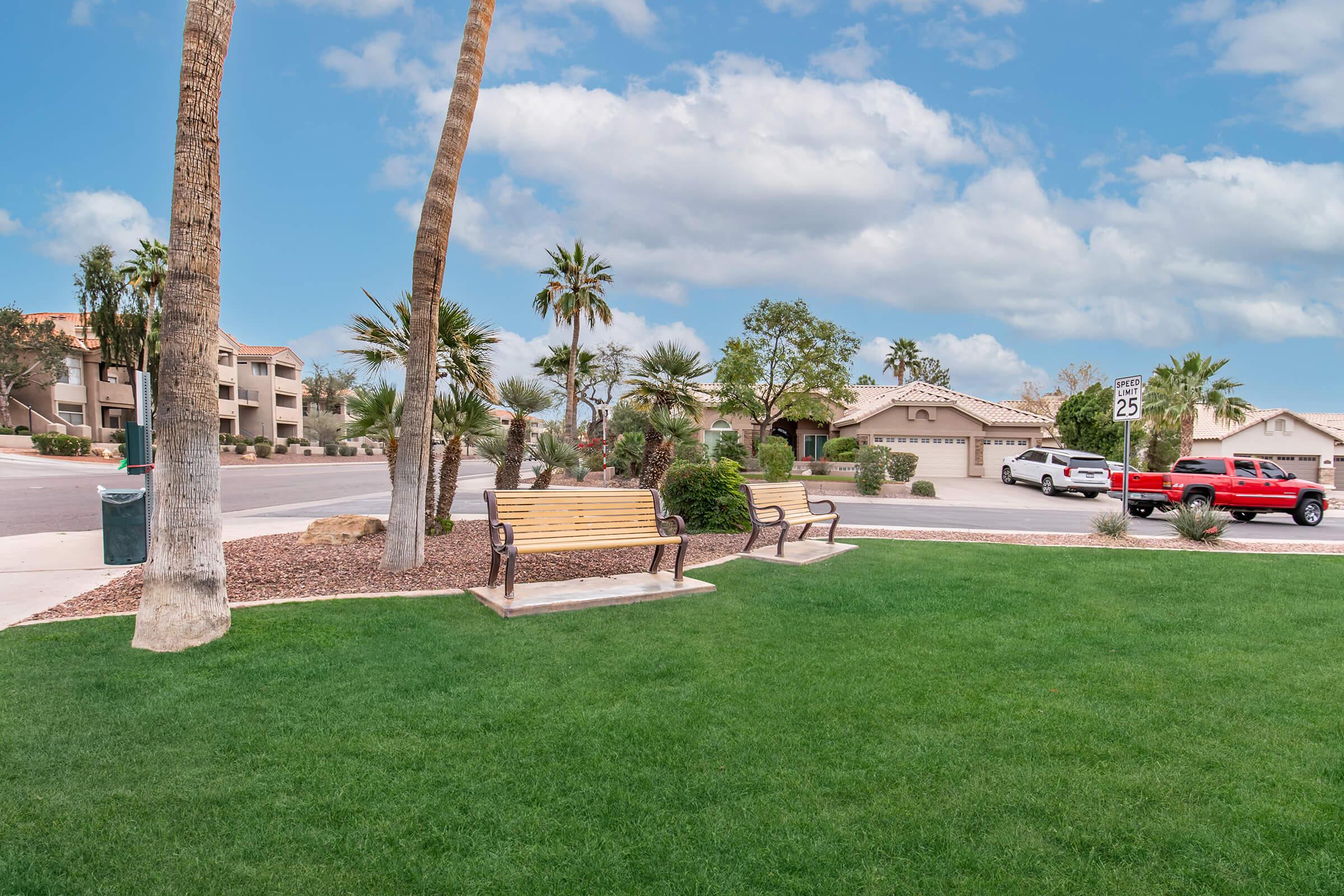 A peaceful neighborhood scene featuring a green lawn with two wooden benches. Surrounding the area are palm trees and other landscaping. In the background, residential buildings are visible along with parked cars and a street sign indicating a 25 mph speed limit. The sky is partly cloudy.