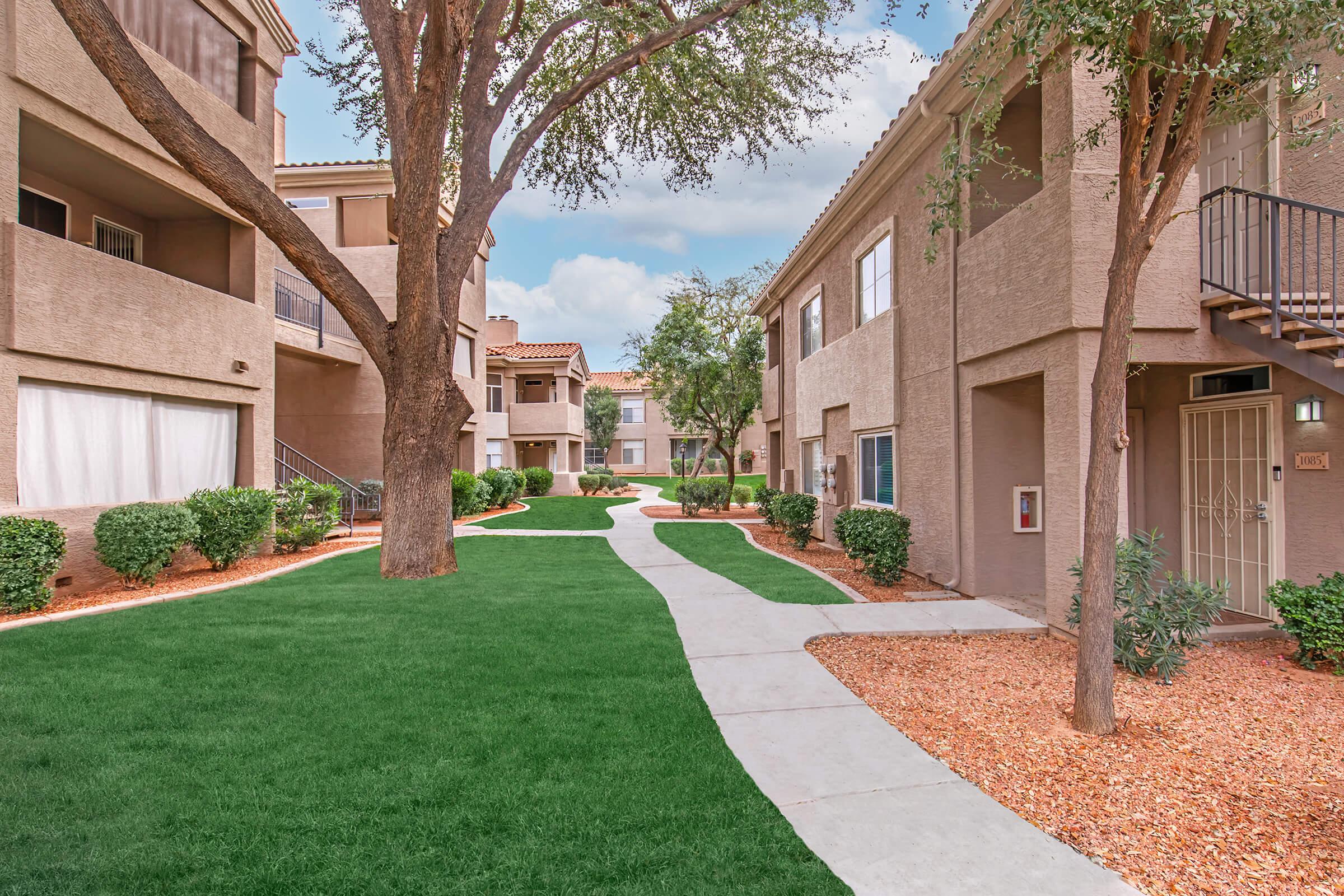 A peaceful residential courtyard featuring green grass, winding pathways, and surrounding buildings with balconies. There are trees and shrubbery enhancing the tranquil atmosphere, making it an inviting space for residents. The sky is clear with a few clouds, adding to the serene setting.